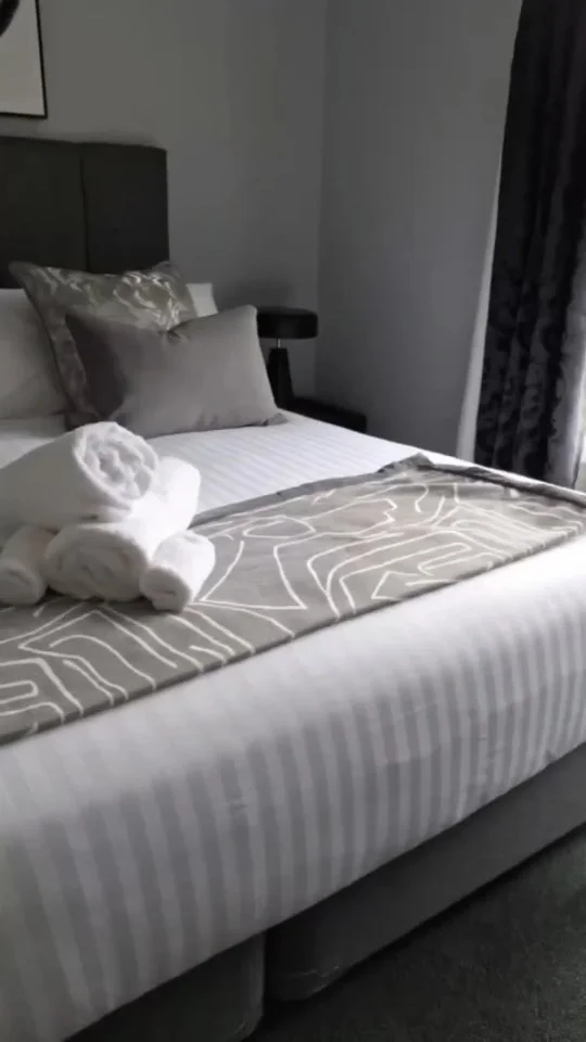 A neatly made bed with white striped sheets, satin pillows, and a patterned beige runner. Towels are rolled and placed on top of the bed.