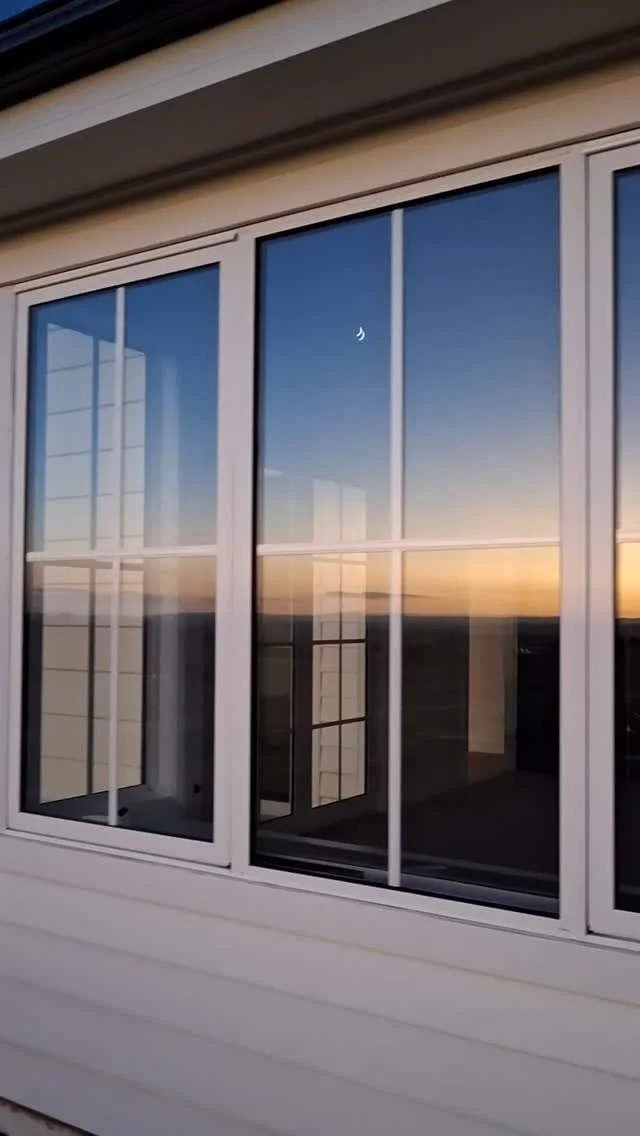 A view through a large window showing a sunset sky with a crescent moon, and the reflection of the interior and exterior of a house.