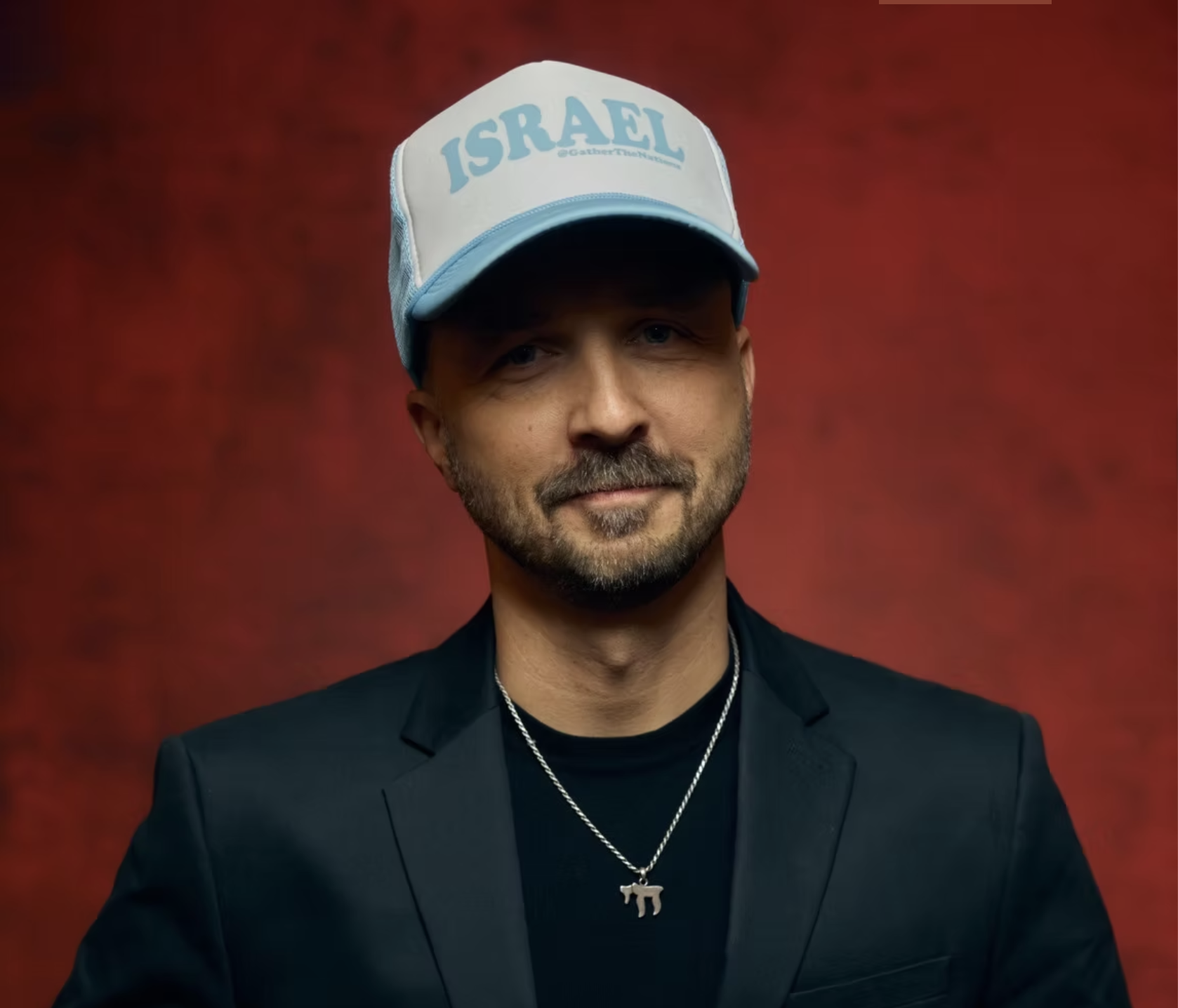 A man is wearing a black blazer, a black shirt, a silver necklace with a pendant, and a white cap with blue accents that reads 'ISRAEL.' He has a beard and mustache, and is standing against a reddish textured background.