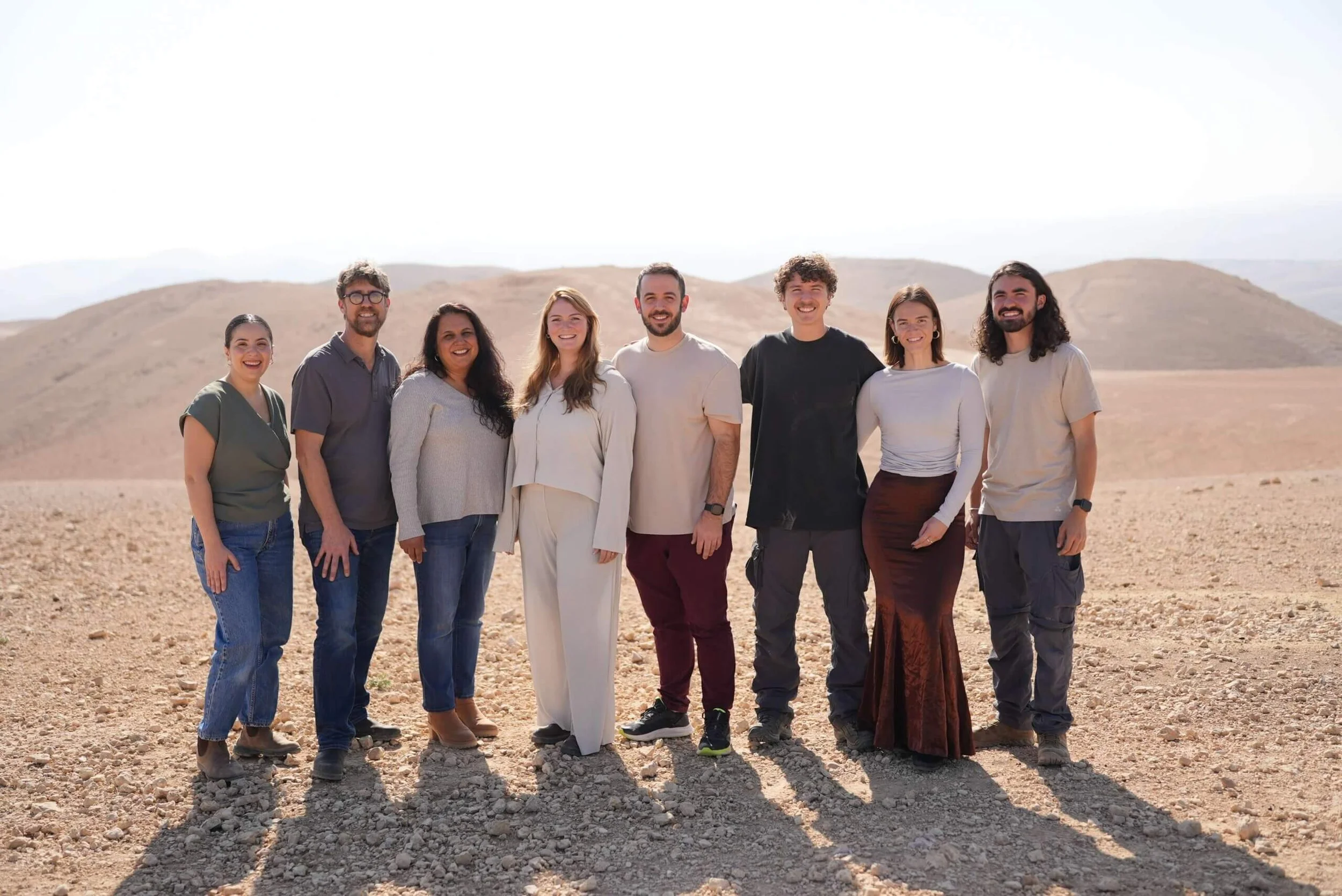 A diverse group of eight people standing in a desert with mountains in the background, smiling and posing for the photo.