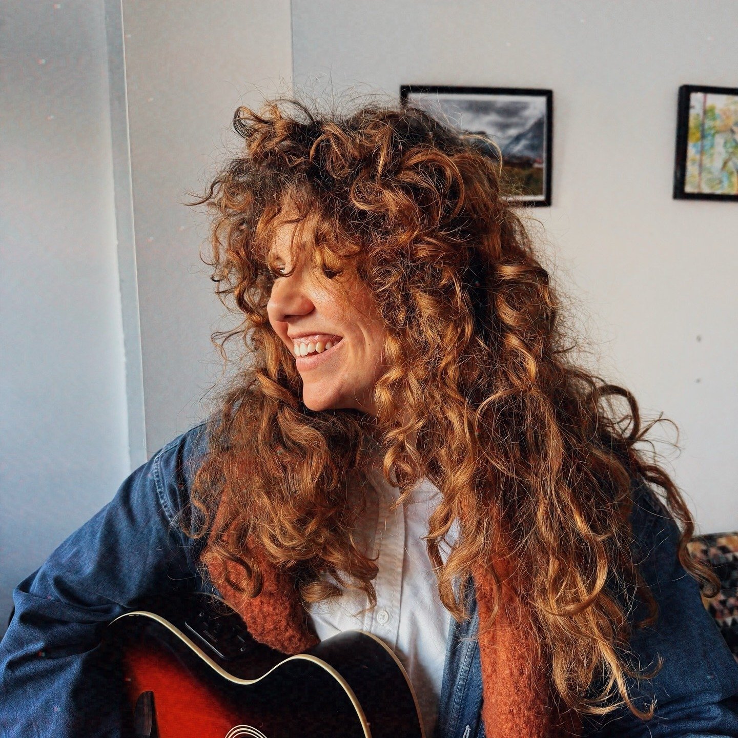 A woman with curly red hair smiling and playing an acoustic guitar, sitting indoors with framed pictures on the wall behind her.
