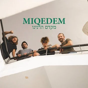 Four people standing on a balcony, looking down at the camera, with a sign that says 'MIQDEM' and text in Hebrew.