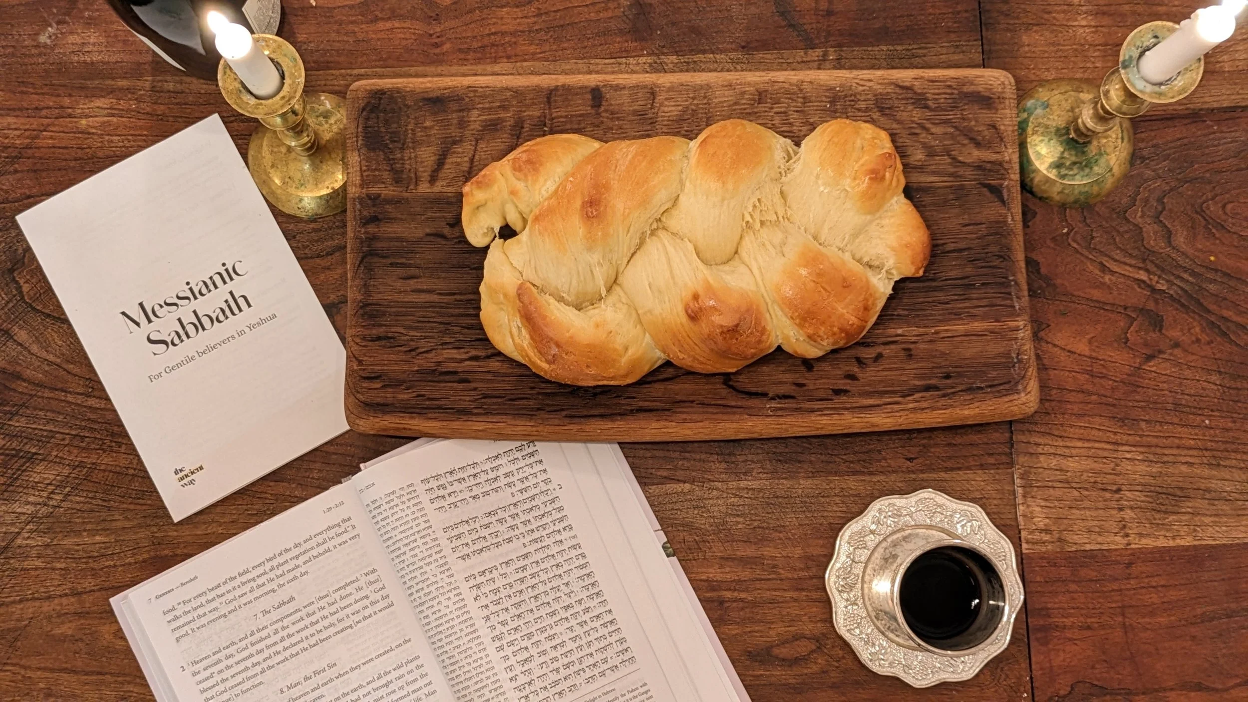 A braided challah bread on a wooden serving board, Bible with Hebrew text, a booklet titled 'Messianic Sabbath,' a silver cup of wine on an ornate coaster, and two candles in brass candlesticks are arranged on a wooden table.