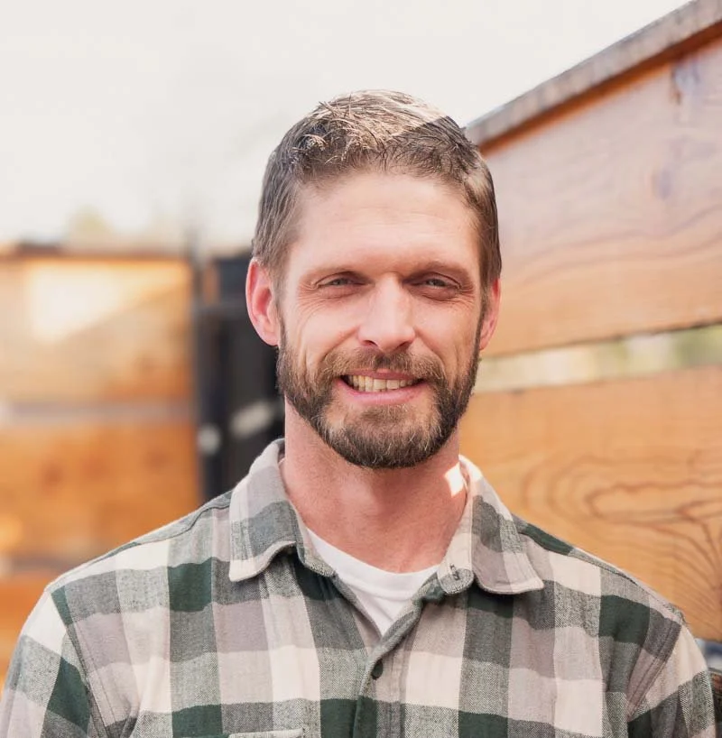 A smiling man with brown hair, a beard, and wearing a plaid shirt, standing outdoors in front of a wooden fence.