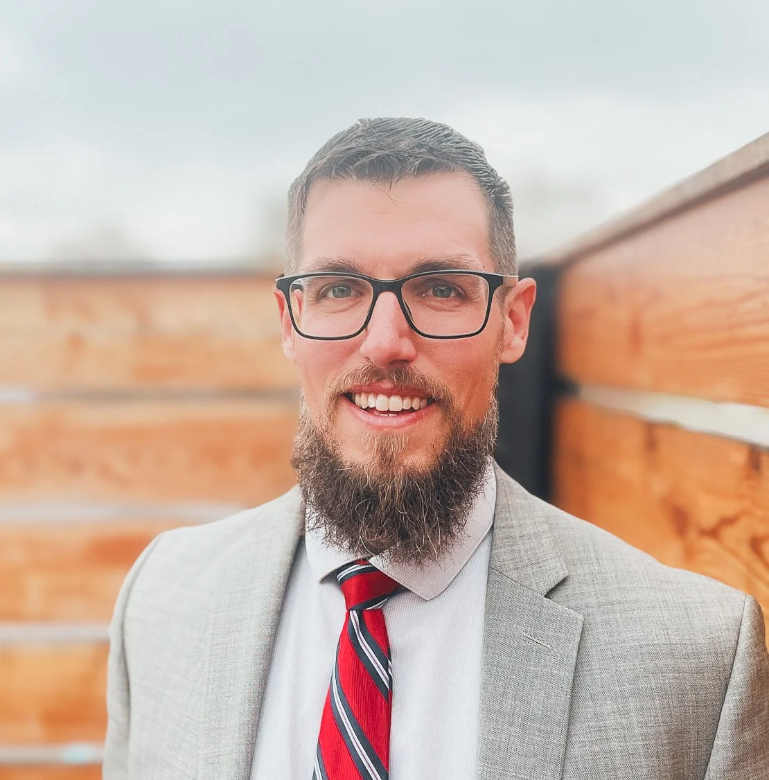 A man with glasses, a beard, and short hair smiling, wearing a light gray suit, white shirt, and red striped tie, standing outdoors in front of a wooden fence.