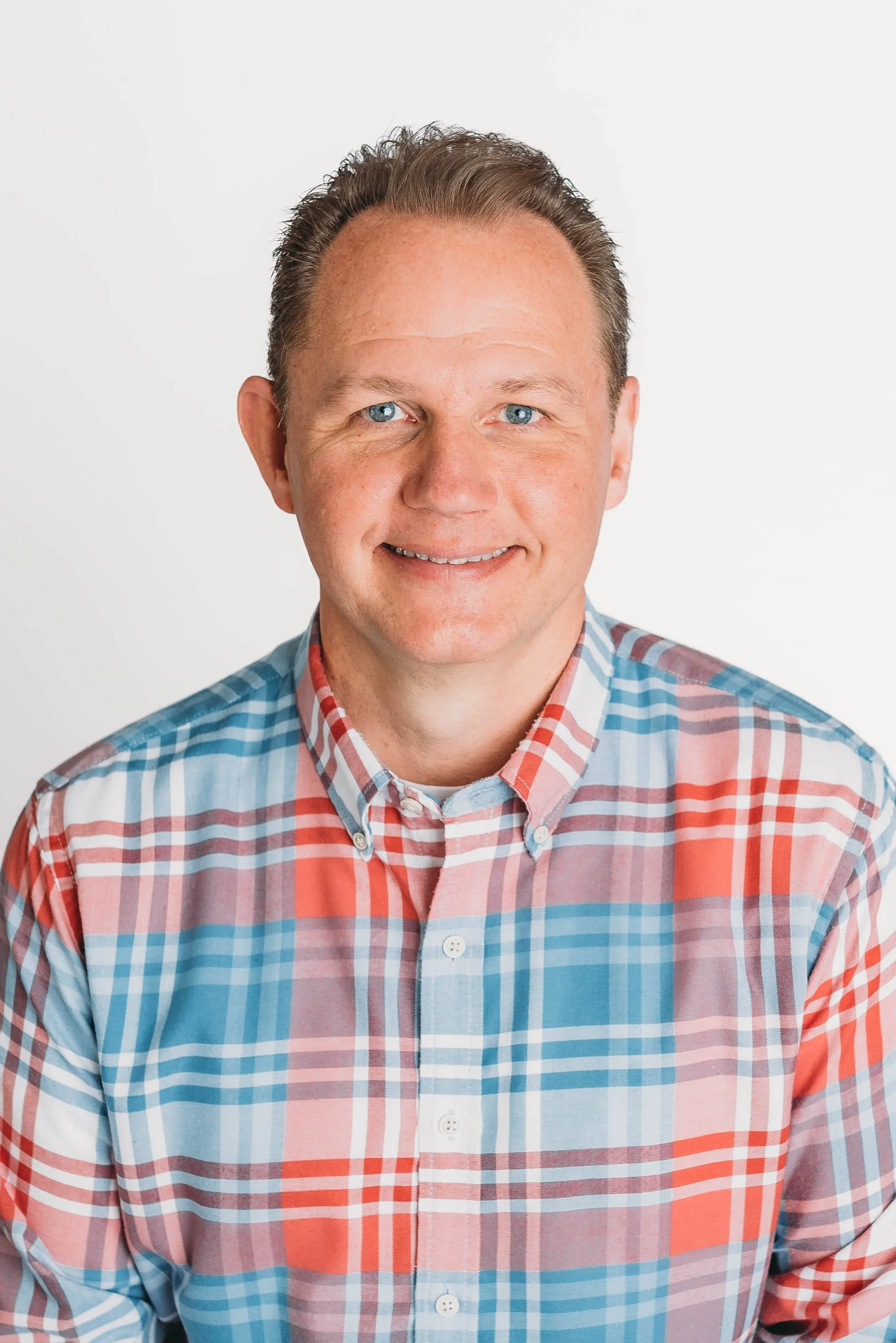 A man with blue eyes and short, brown hair smiling at the camera, wearing a colorful plaid button-up shirt against a plain white background.