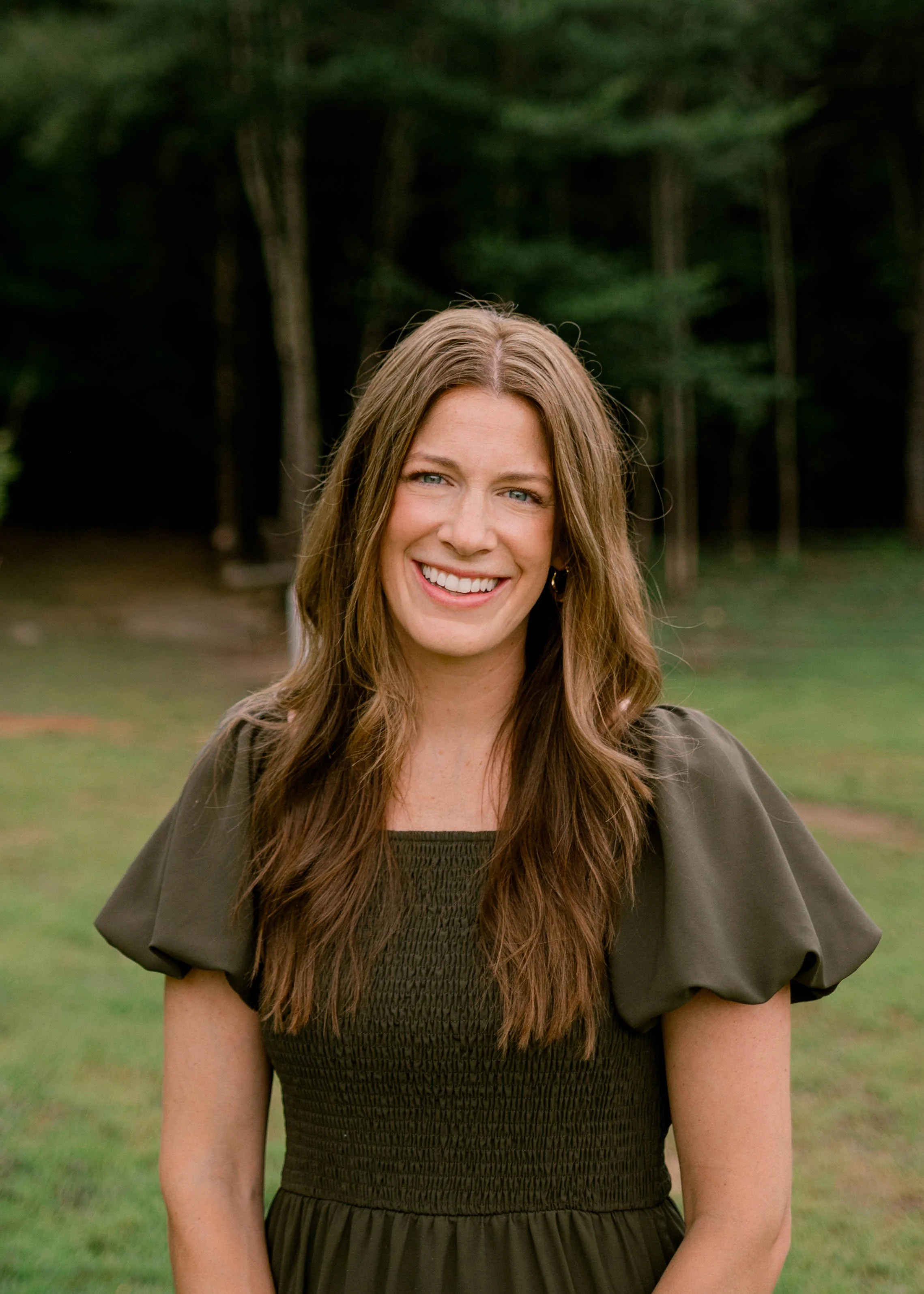 A woman with shoulder-length brown hair wearing a denim jacket and striped shirt, smiling in front of a window with trees outside.