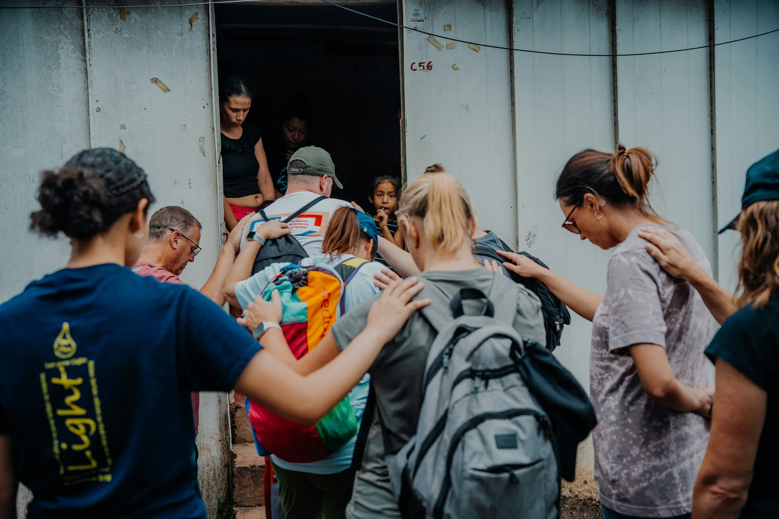 A group of people praying with their heads bowed and hands on each other's shoulders at the entrance of a building with metal doors.