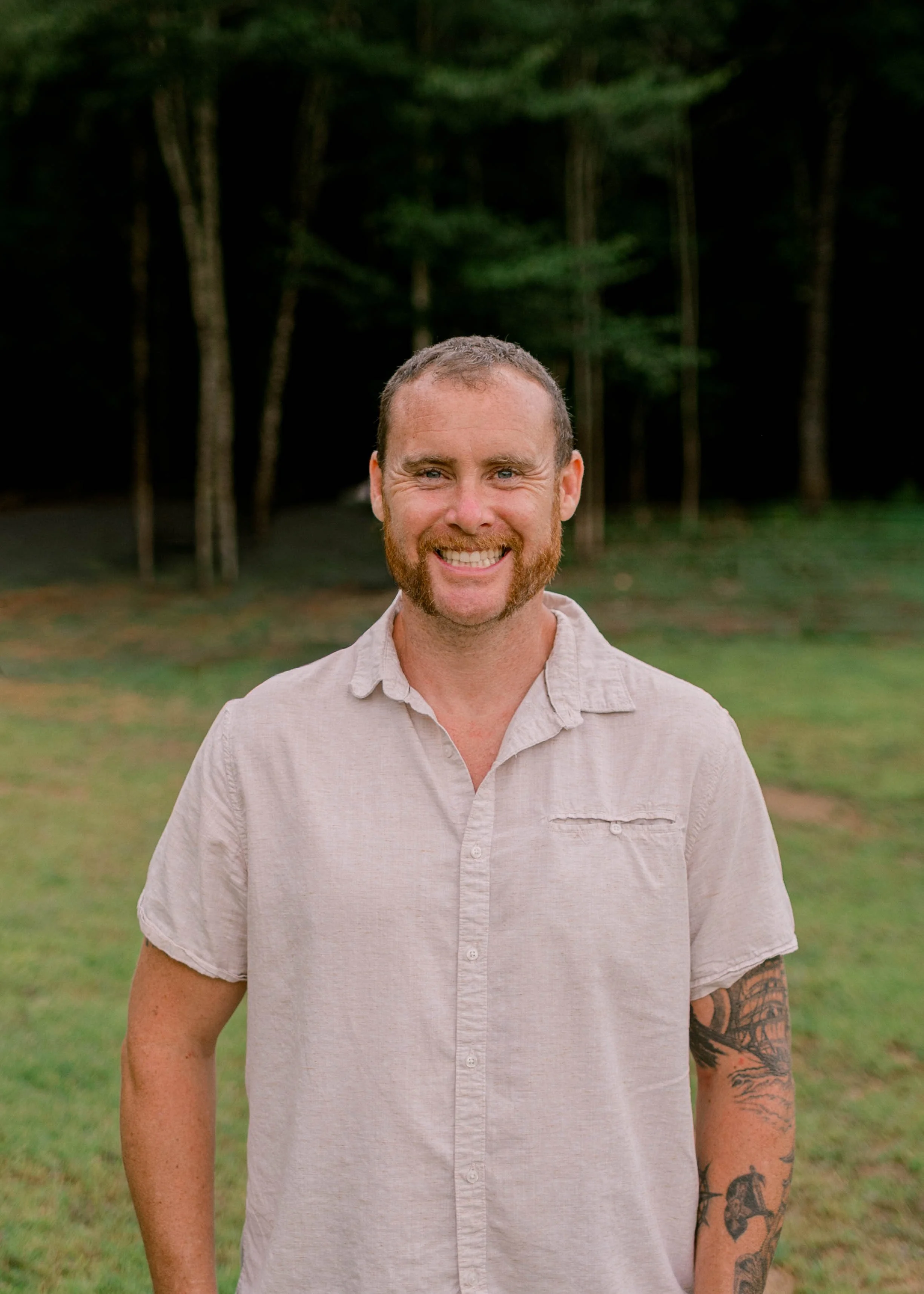 A man with a beard and mustache wearing a teal hoodie, smiling outdoors with green foliage in the background.