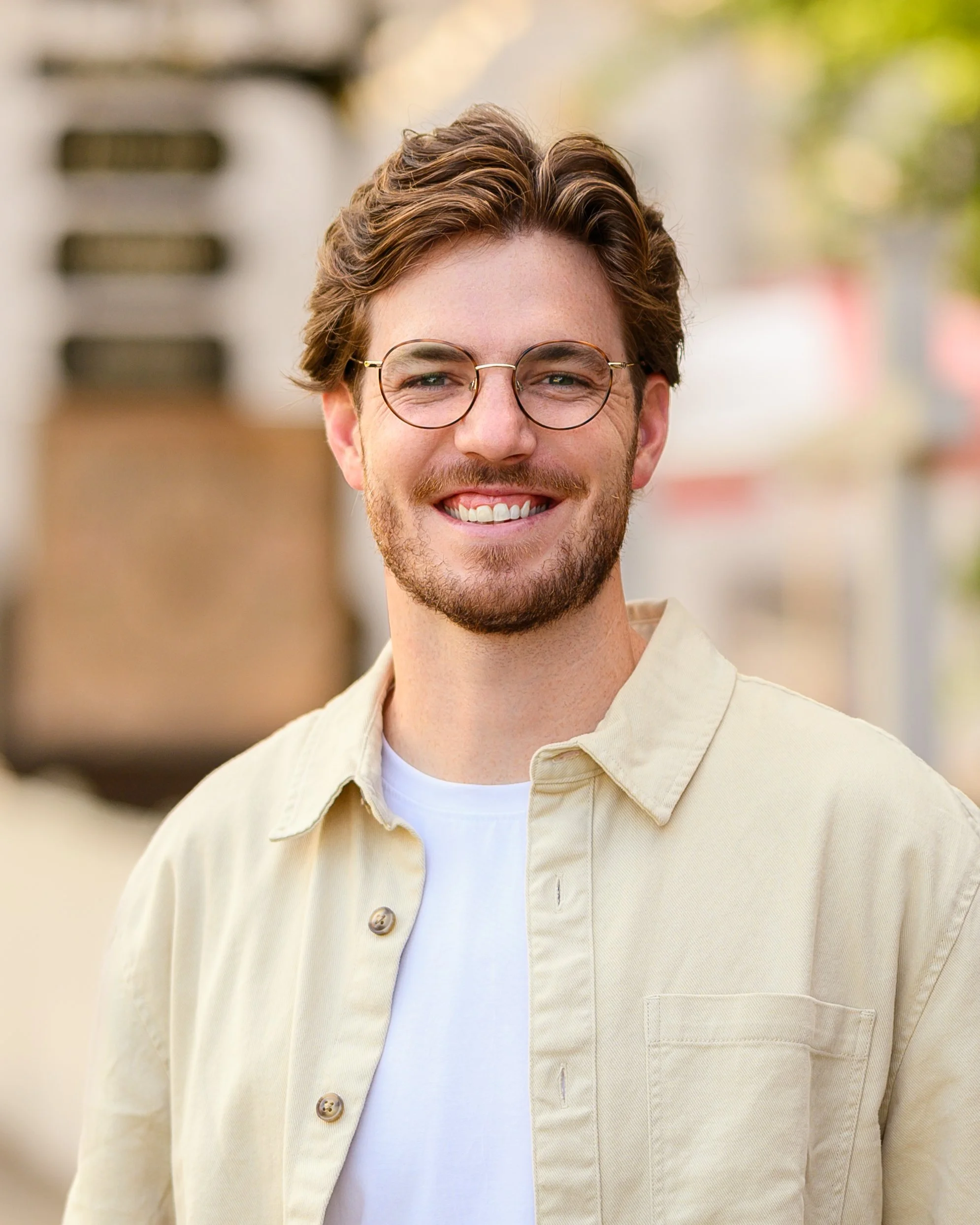 Smiling young man with glasses, beard, and wavy hair standing outdoors, blurred background with trees and buildings.