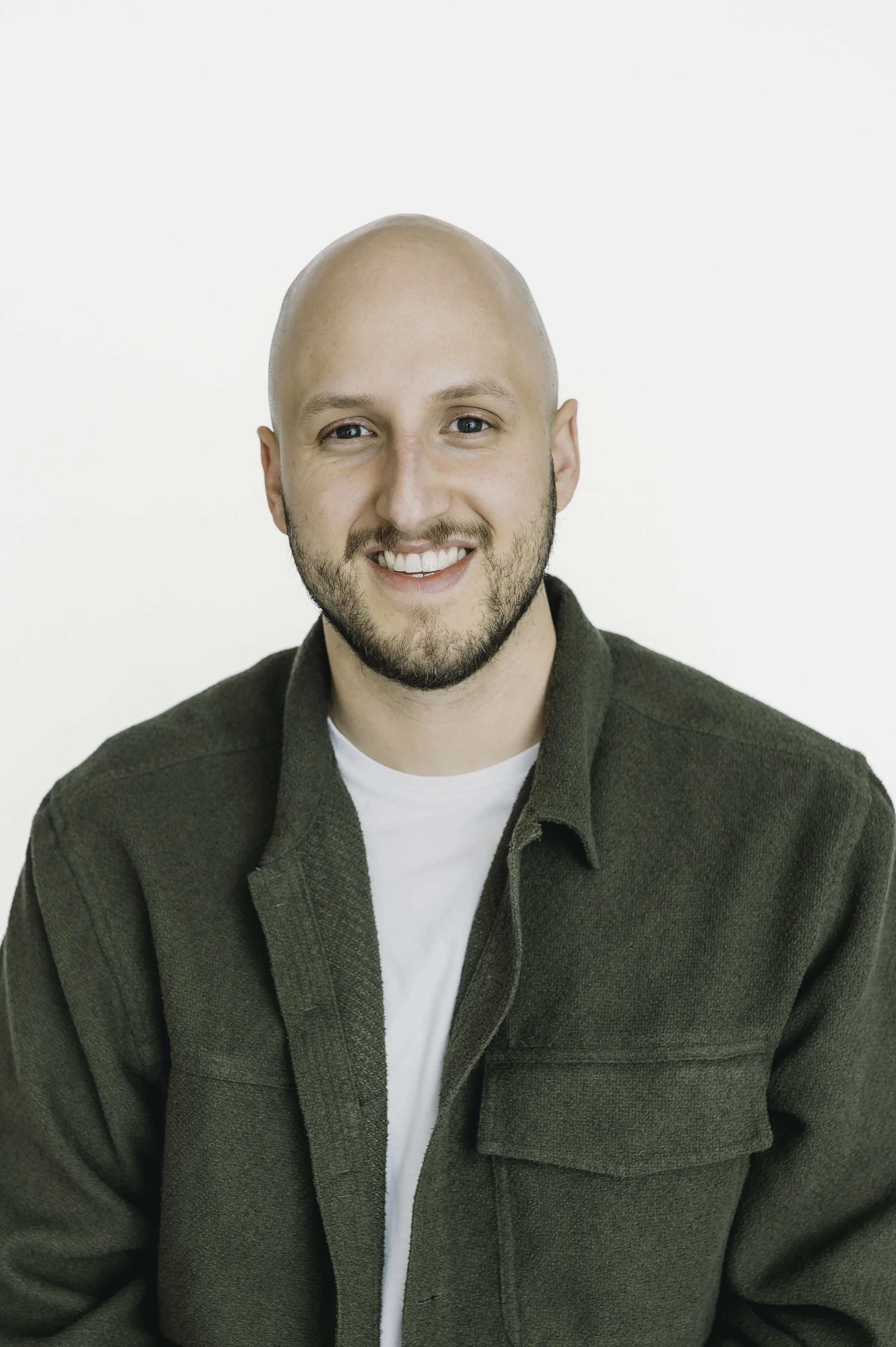 A smiling man with a clean-shaven head and beard, wearing a dark green jacket over a white shirt, against a plain white background.