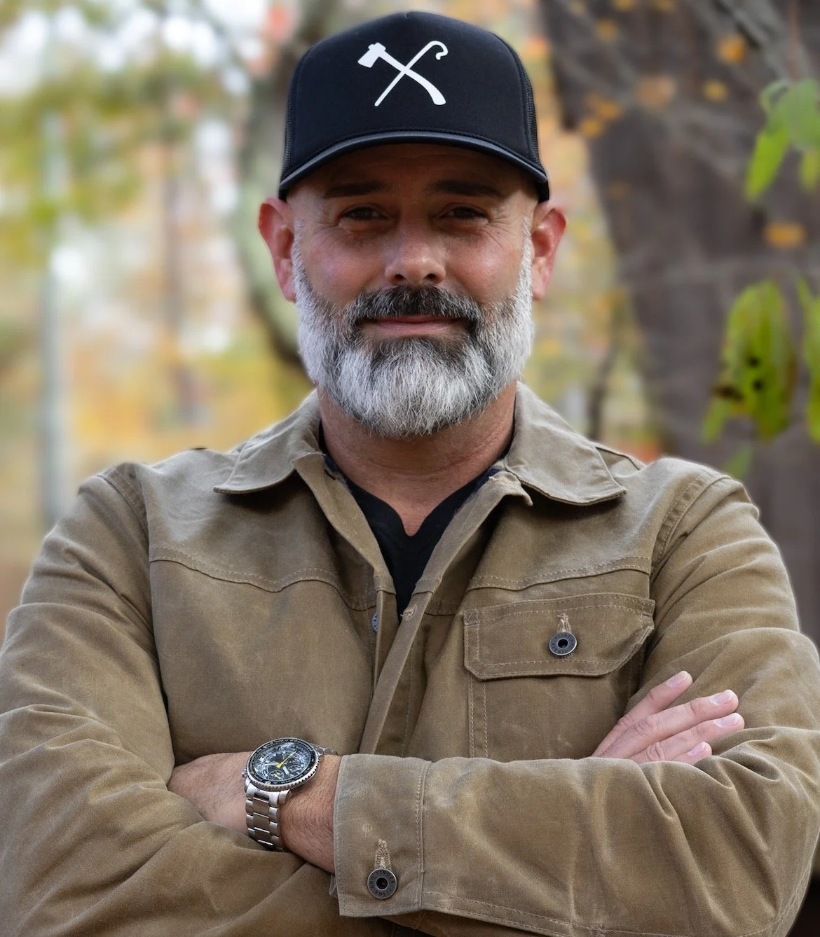 A man with a gray beard and mustache, wearing a black cap with a hammer and pickaxe symbol, brown jacket, and silver watch, standing outdoors with blurred trees and fall foliage in the background.