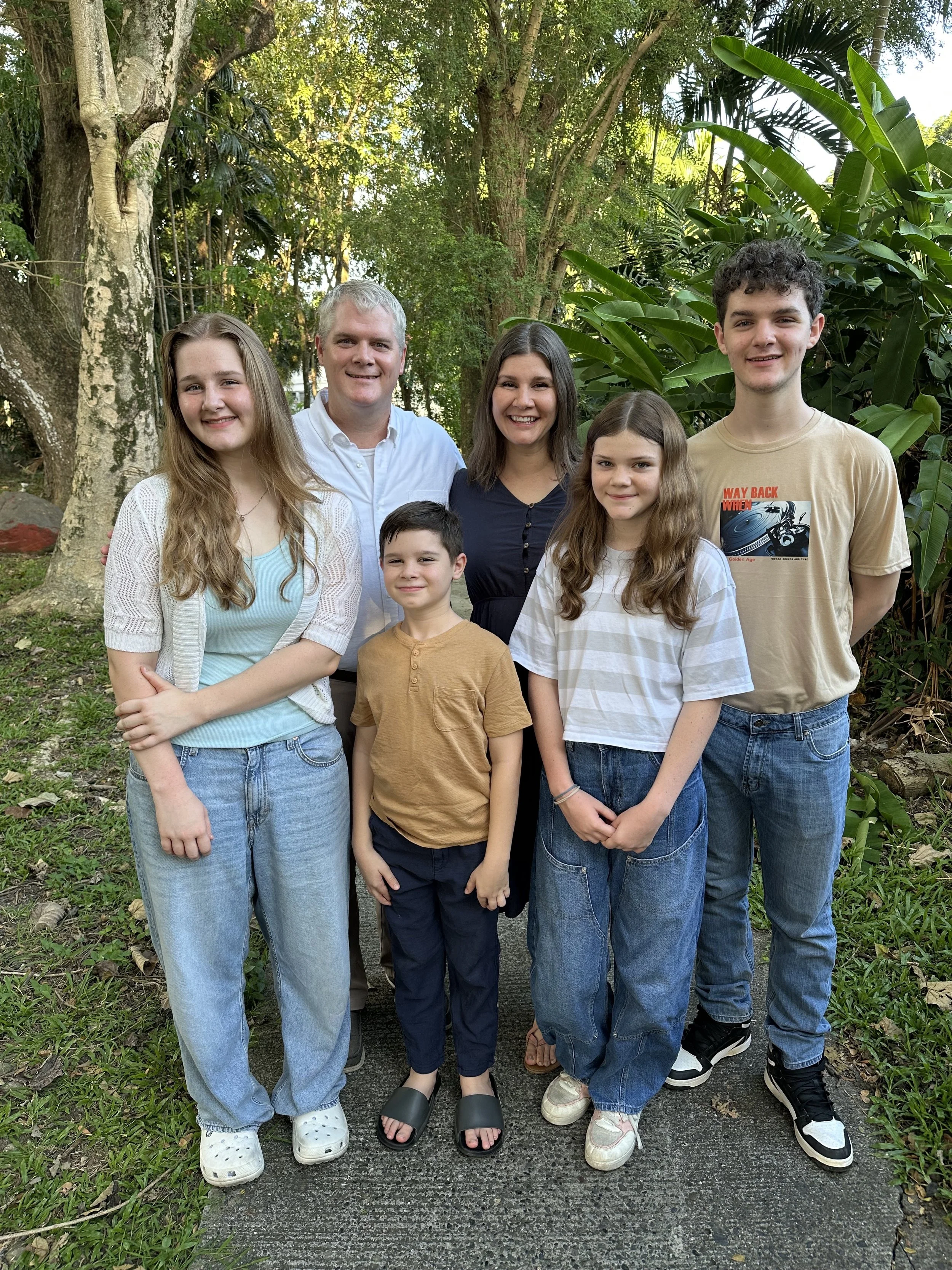 A family of seven posing outdoors on a pathway surrounded by trees and lush greenery, smiling at the camera.