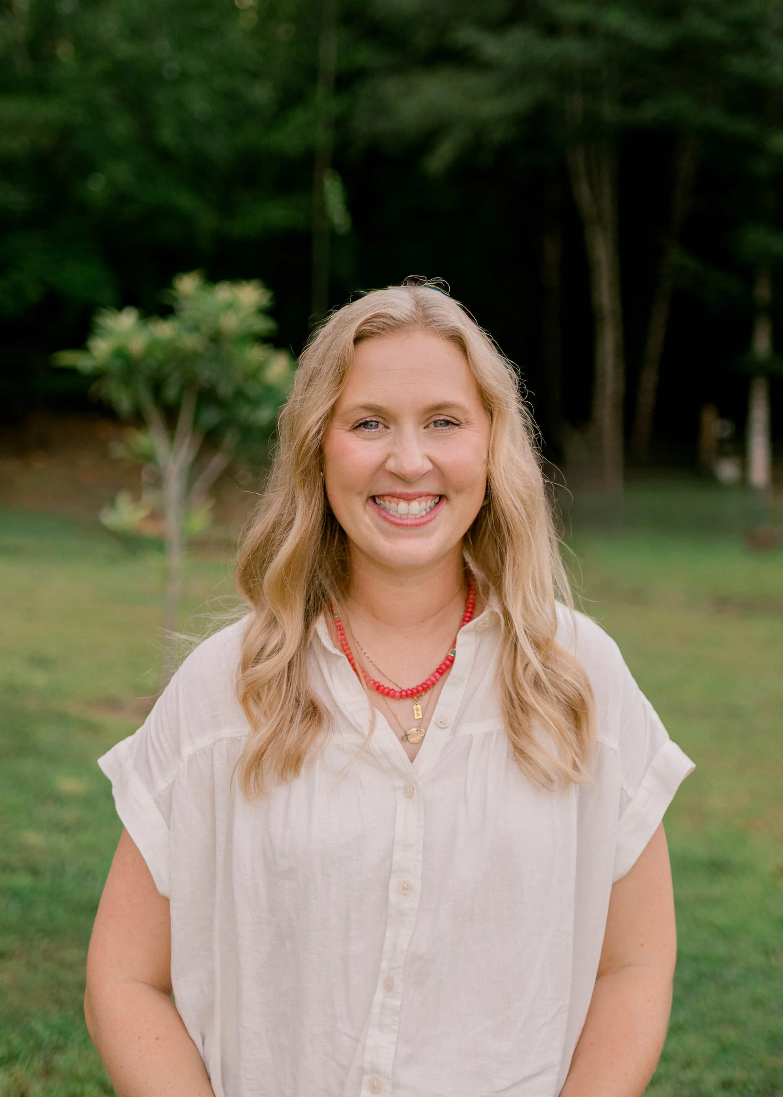 A young woman with long, wavy blonde hair, smiling in front of a plain light beige background. She is wearing a white, textured, short-sleeved dress and a delicate necklace.