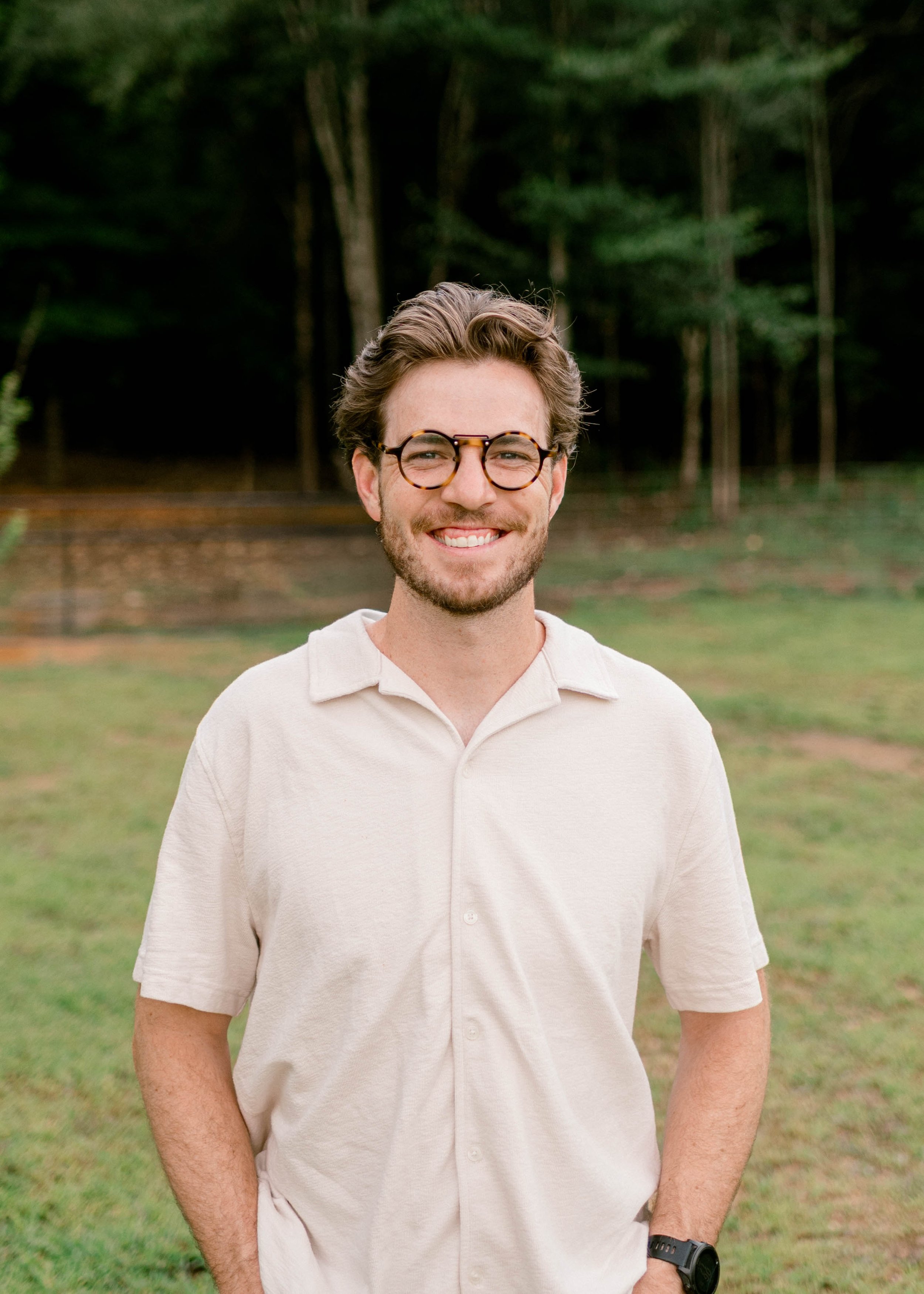 Smiling young man with glasses, beard, and wavy hair standing outdoors, blurred background with trees and buildings.