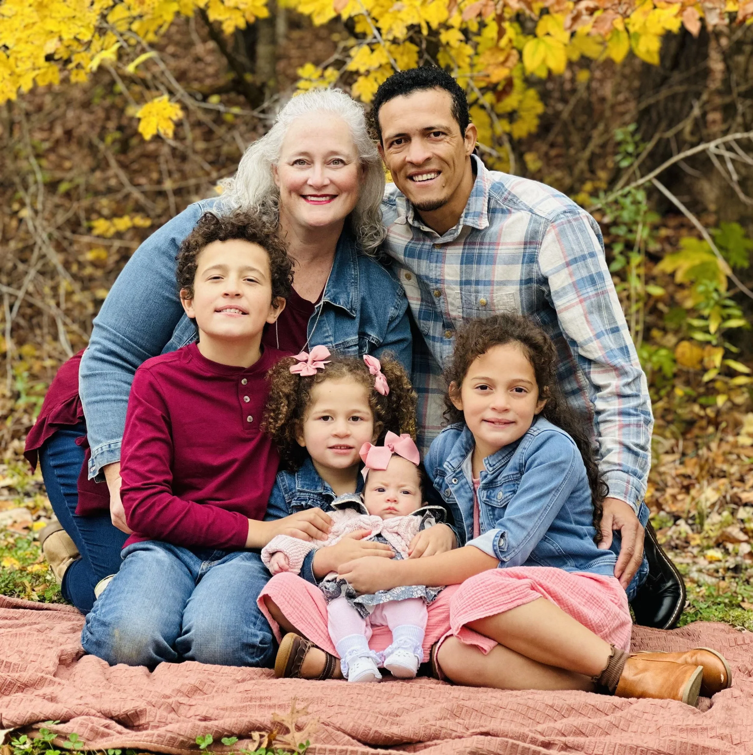A multigenerational family posing outdoors in autumn, sitting on a blanket with fall foliage in the background.