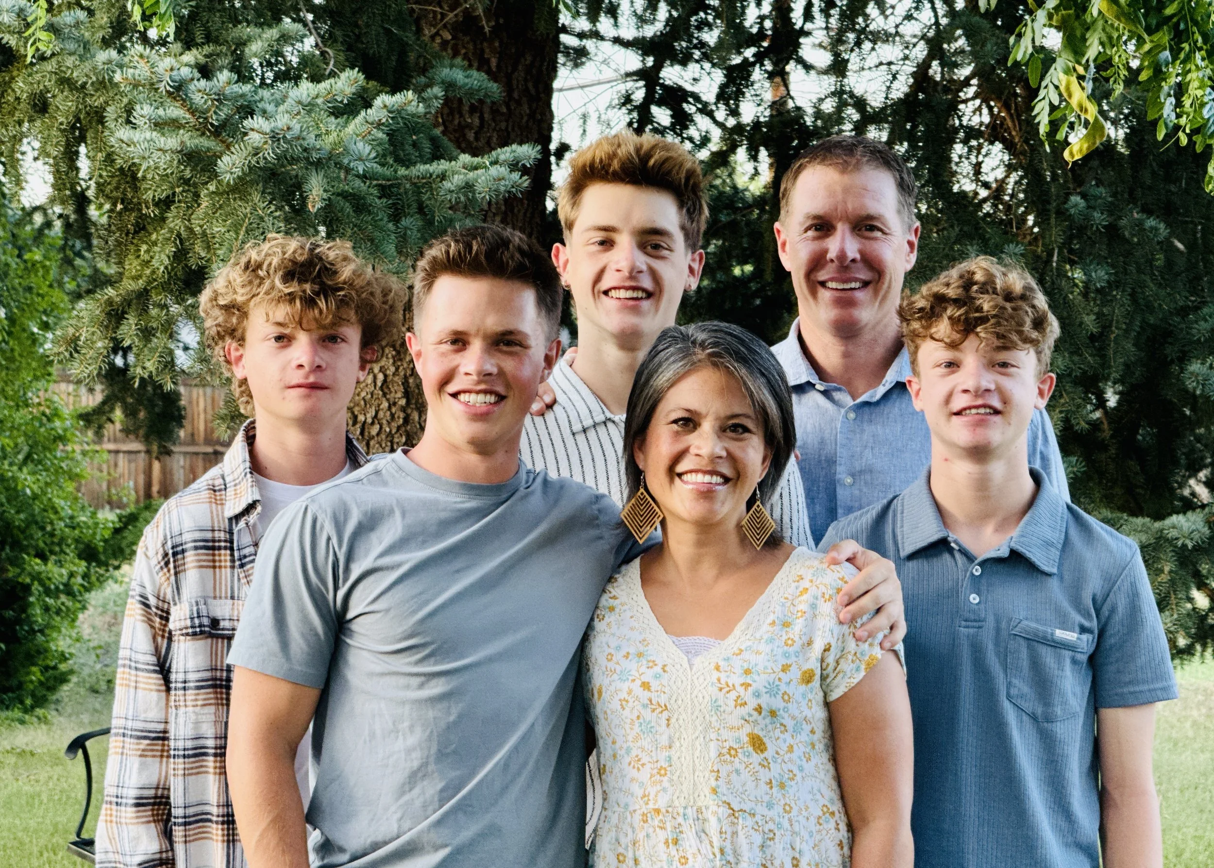 A smiling family of six people, three children, two adults, and one elderly woman, standing outdoors in front of trees and greenery.