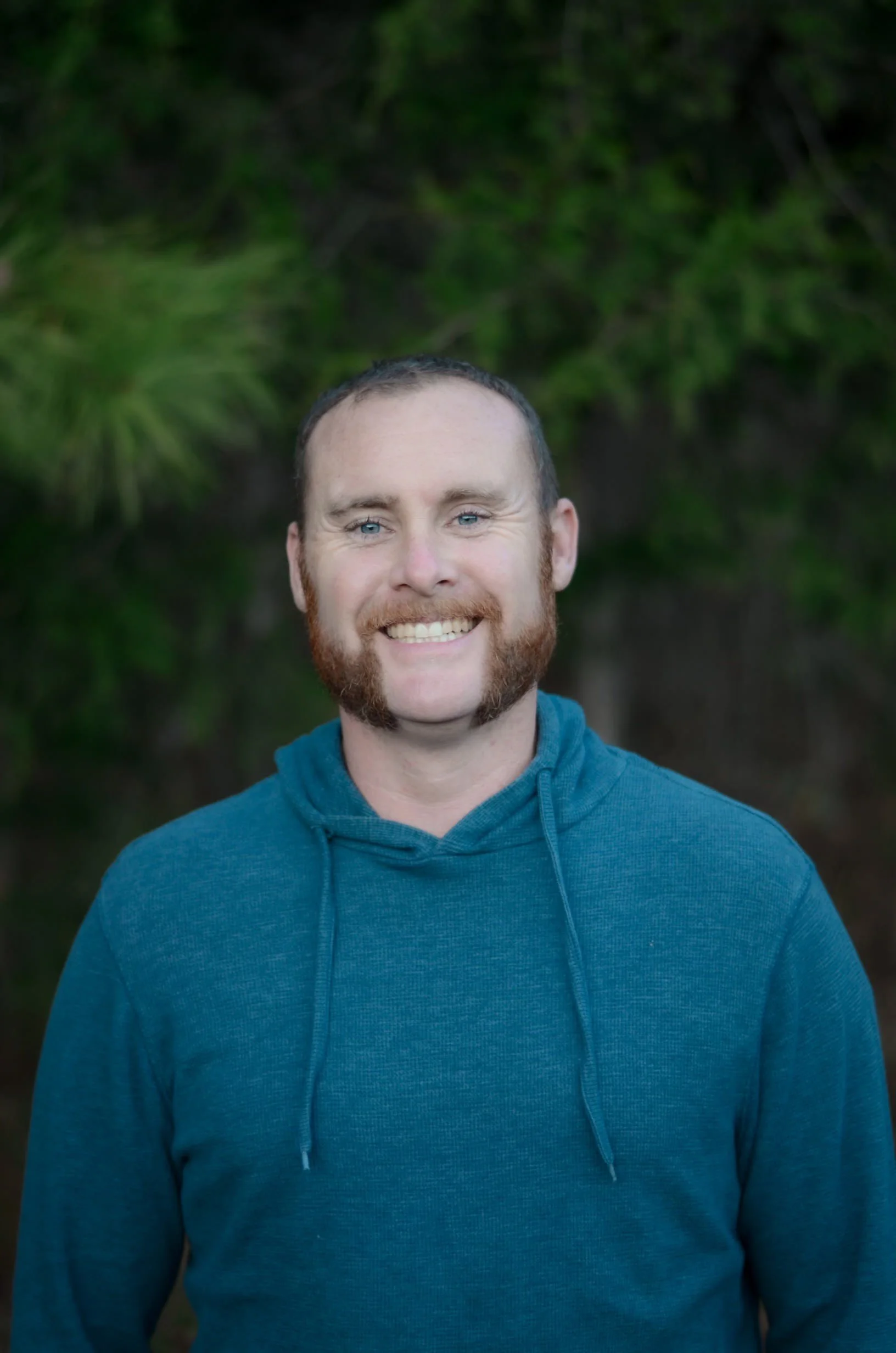 A man with a beard and mustache wearing a teal hoodie, smiling outdoors with green foliage in the background.
