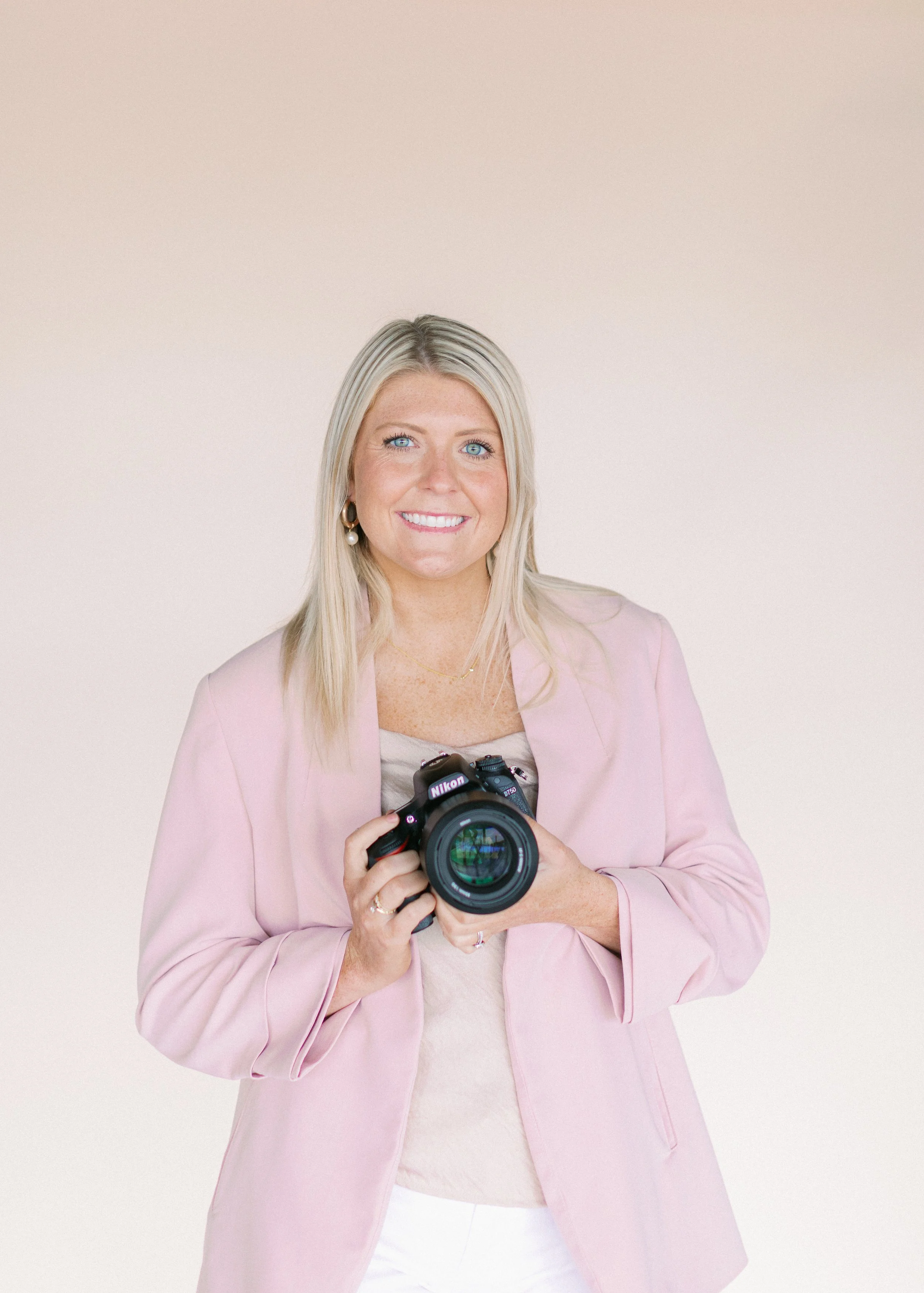 A woman with blonde hair, blue eyes, wearing a pink blazer and earrings, holding a Nikon camera, smiling, against a light neutral background.