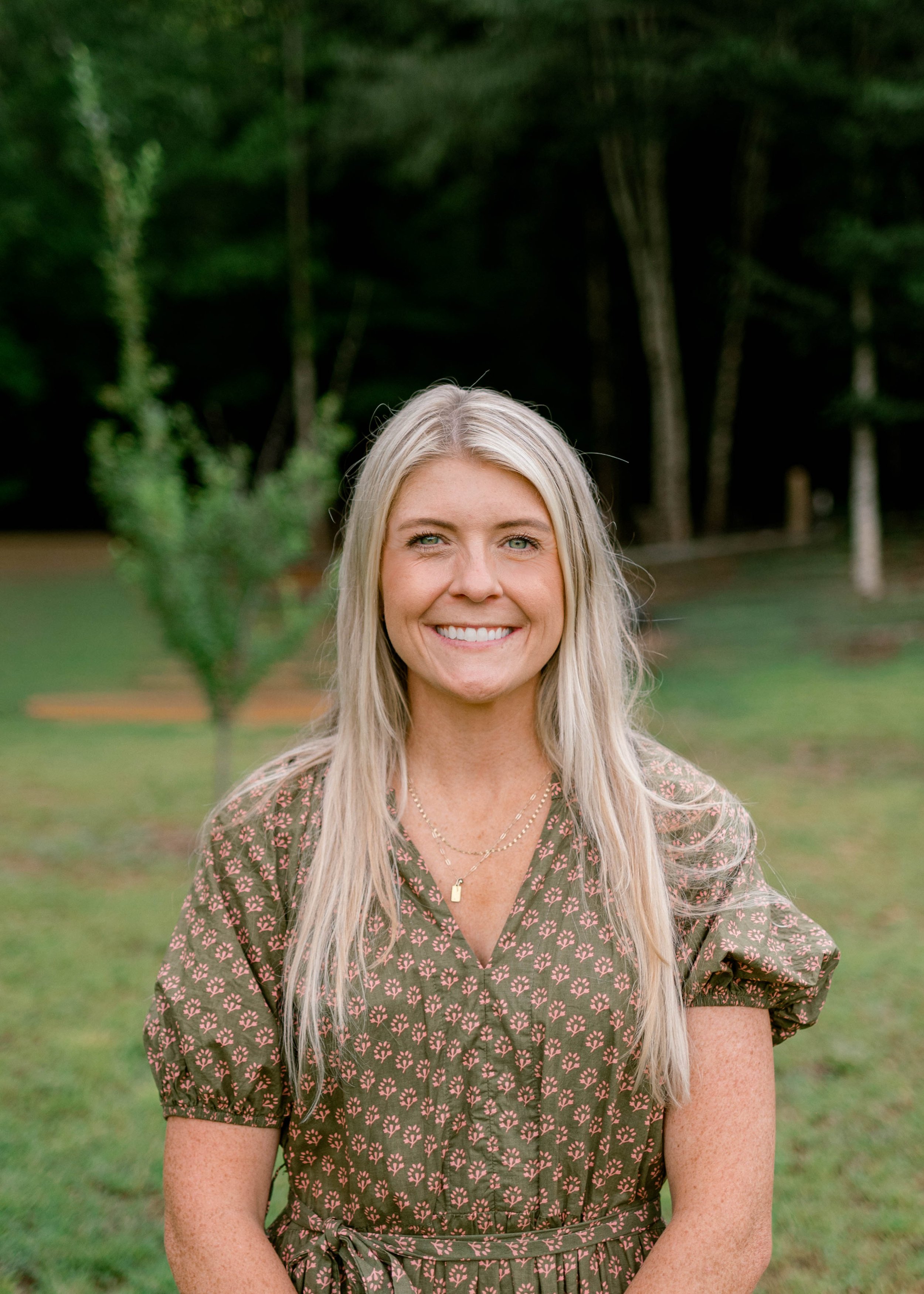 A woman with blonde hair, blue eyes, wearing a pink blazer and earrings, holding a Nikon camera, smiling, against a light neutral background.