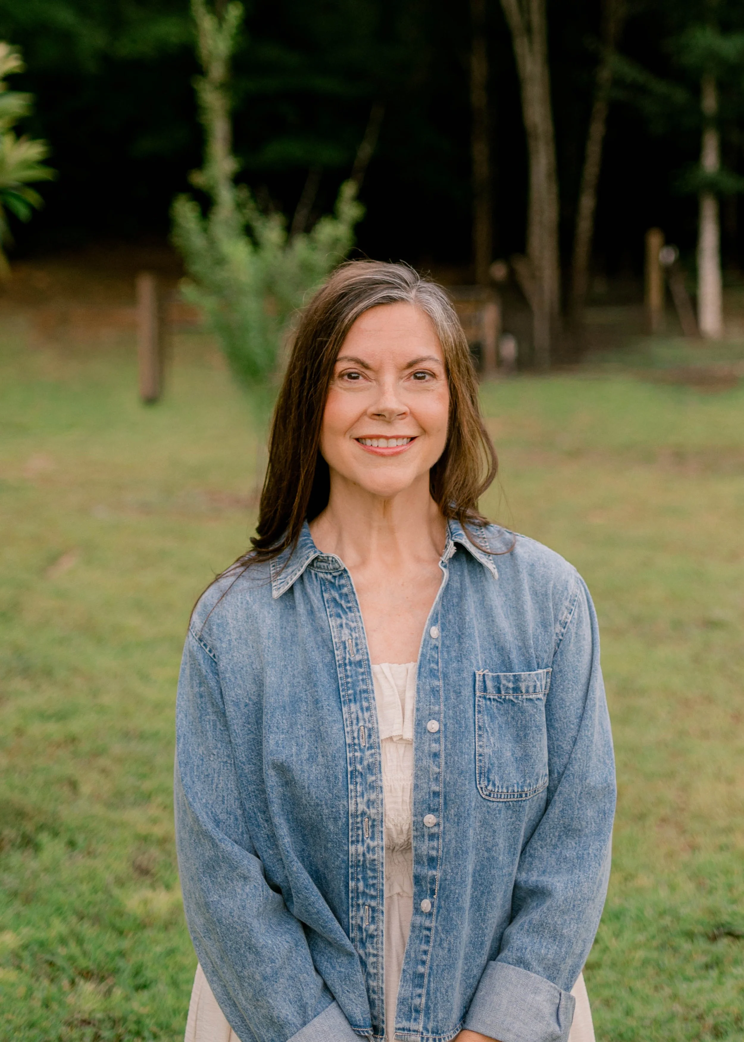 A woman with long brown hair smiling, wearing a blue floral blouse and colorful earrings, against a white background.