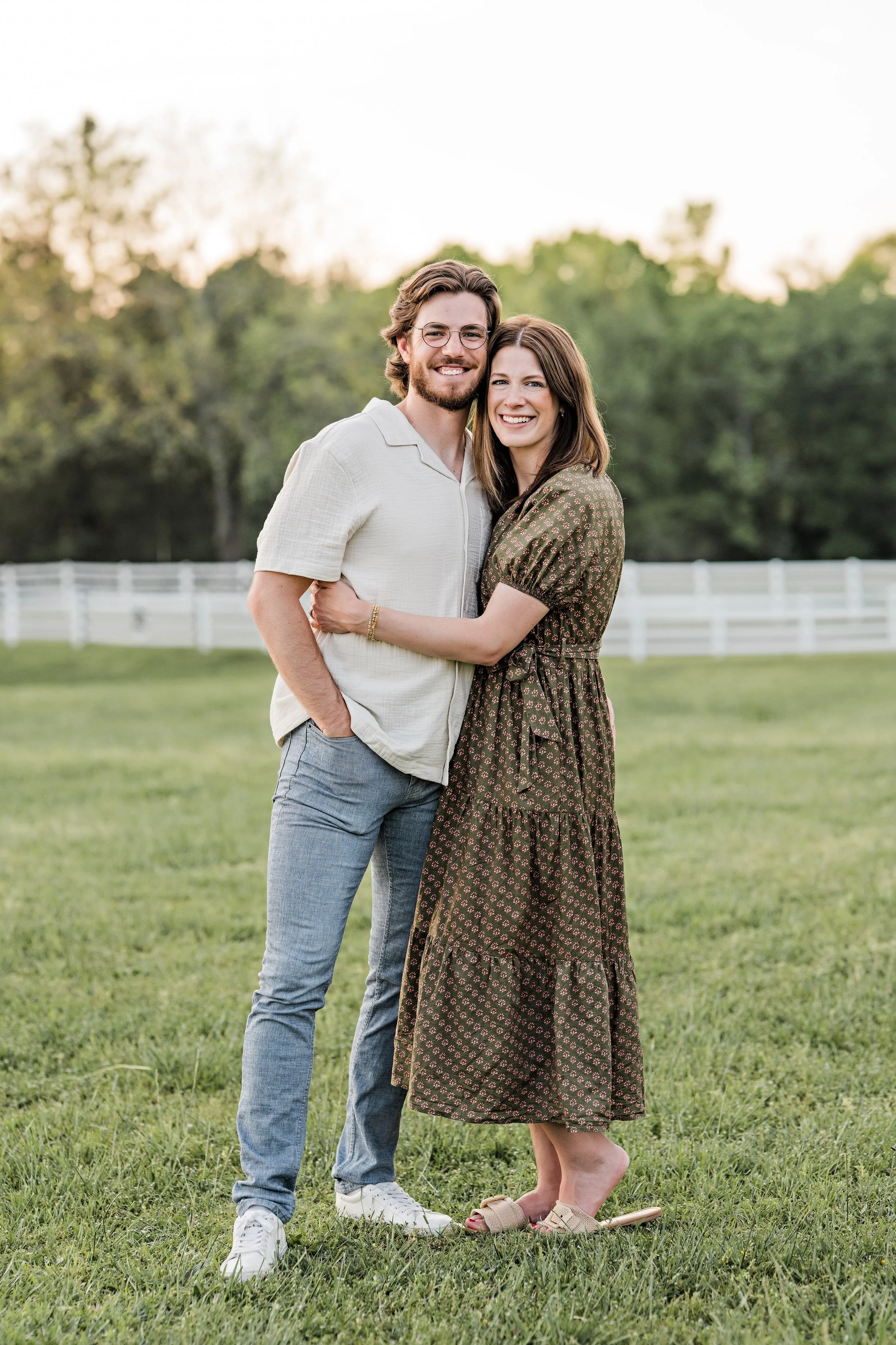 A smiling couple, a man with glasses and a woman with brown hair, standing close together in a grassy field with a white fence and trees in the background during sunset.