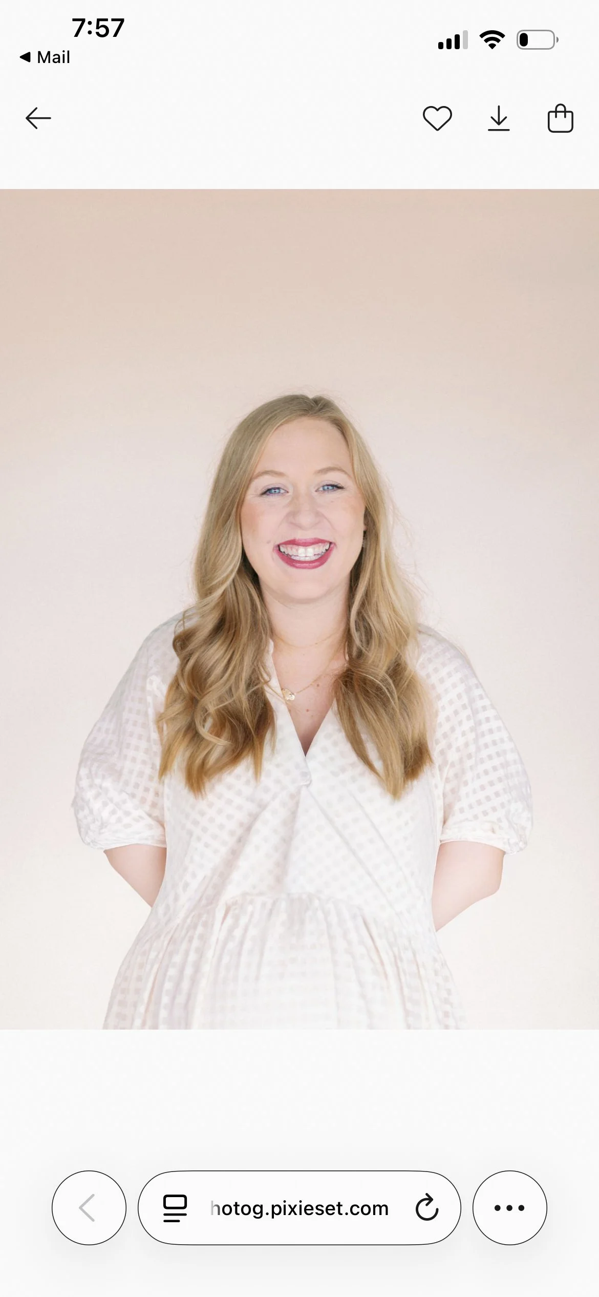 A young woman with long, wavy blonde hair, smiling in front of a plain light beige background. She is wearing a white, textured, short-sleeved dress and a delicate necklace.