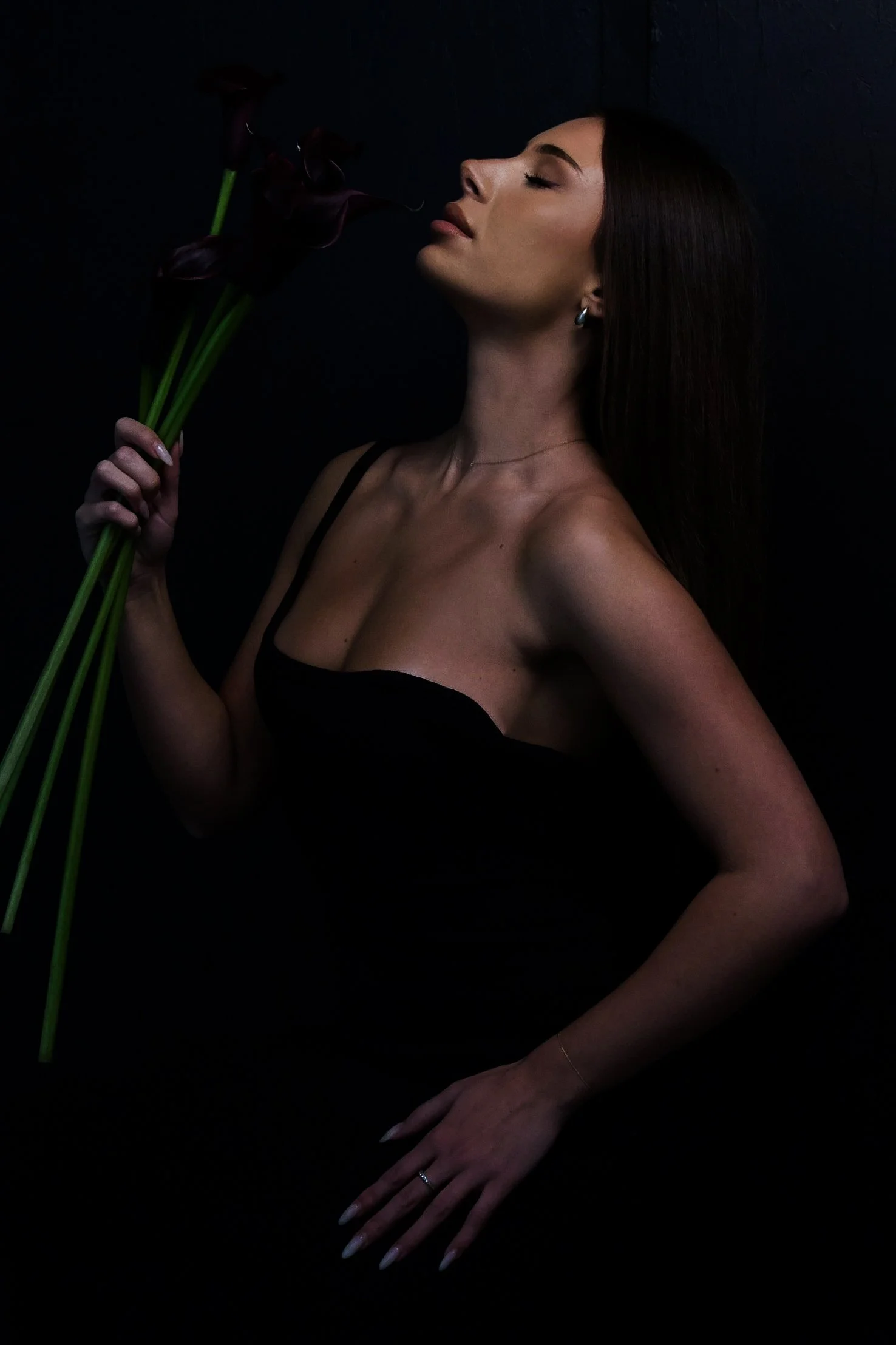 A woman with long brown hair and earrings holds dark flowers close to her face against a dark background. She wears a black strap dress and has a delicate gold necklace and ring.