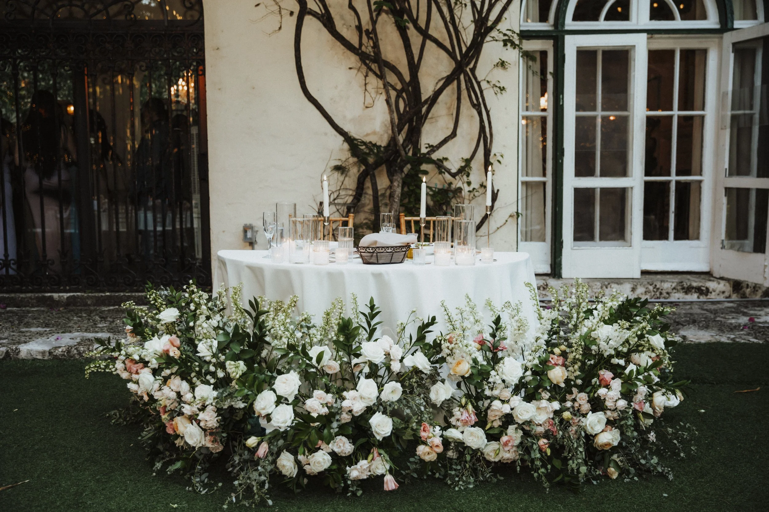 Elegant outdoor table setup with white tablecloth, candles, glassware, and floral centerpiece featuring white and pink flowers, against a backdrop of a building with vines and glass doors.