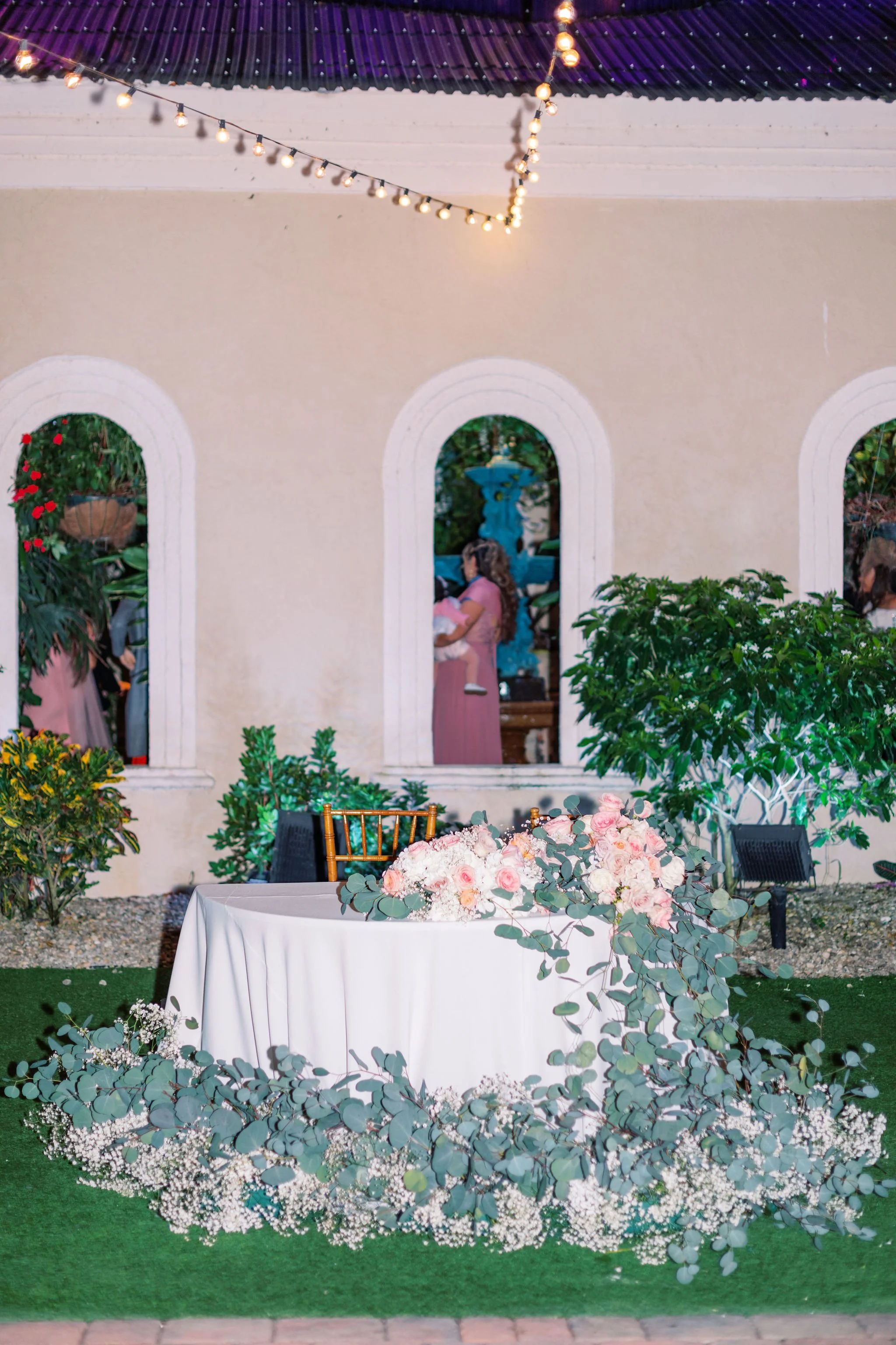 Outdoor wedding reception setup with a round table decorated with pink and white flowers and greenery, string lights overhead, and arched windows in the background.