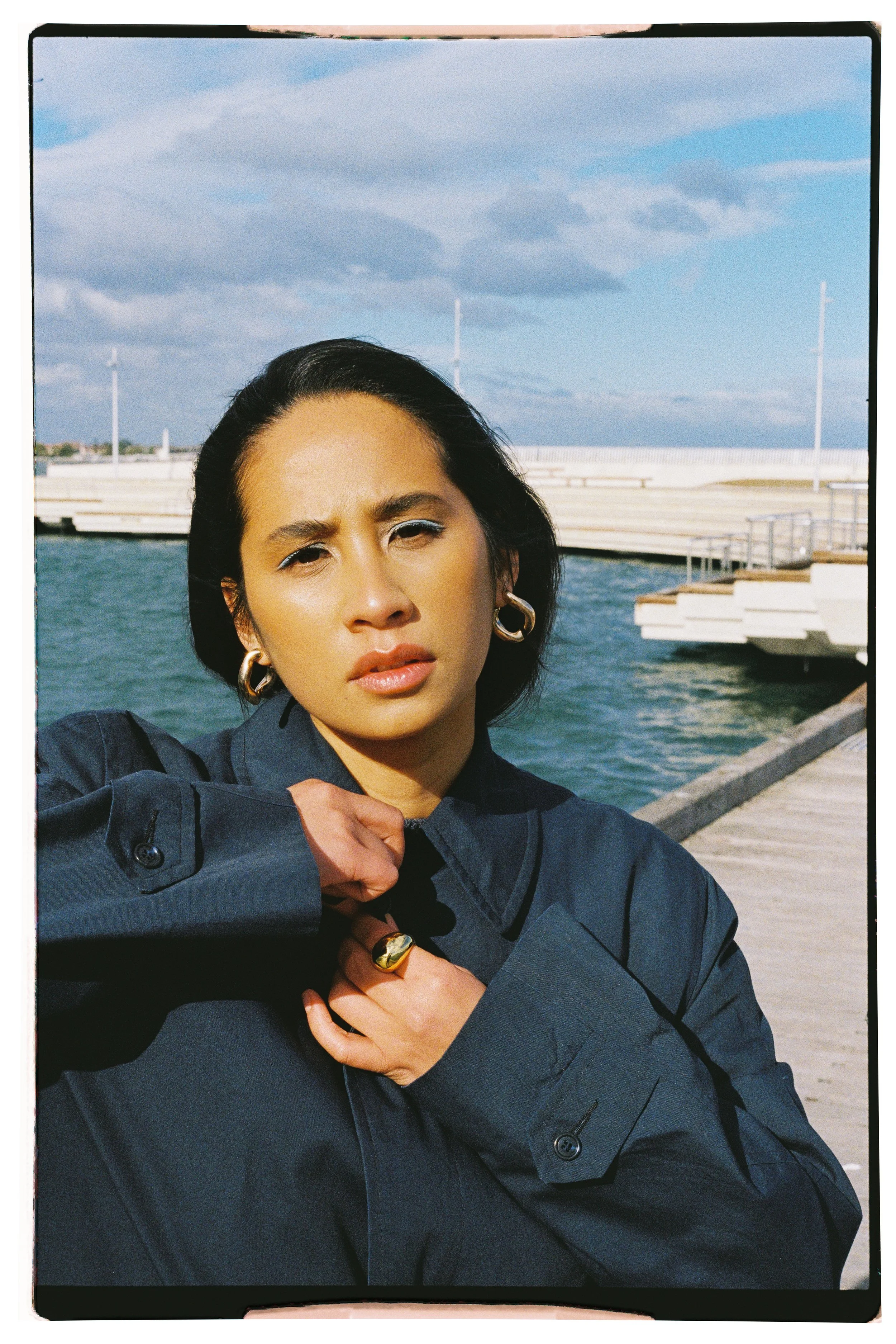 A woman with dark hair and large gold earrings taking a selfie near a body of water with a dock and cloudy sky in the background, wearing a dark jacket.