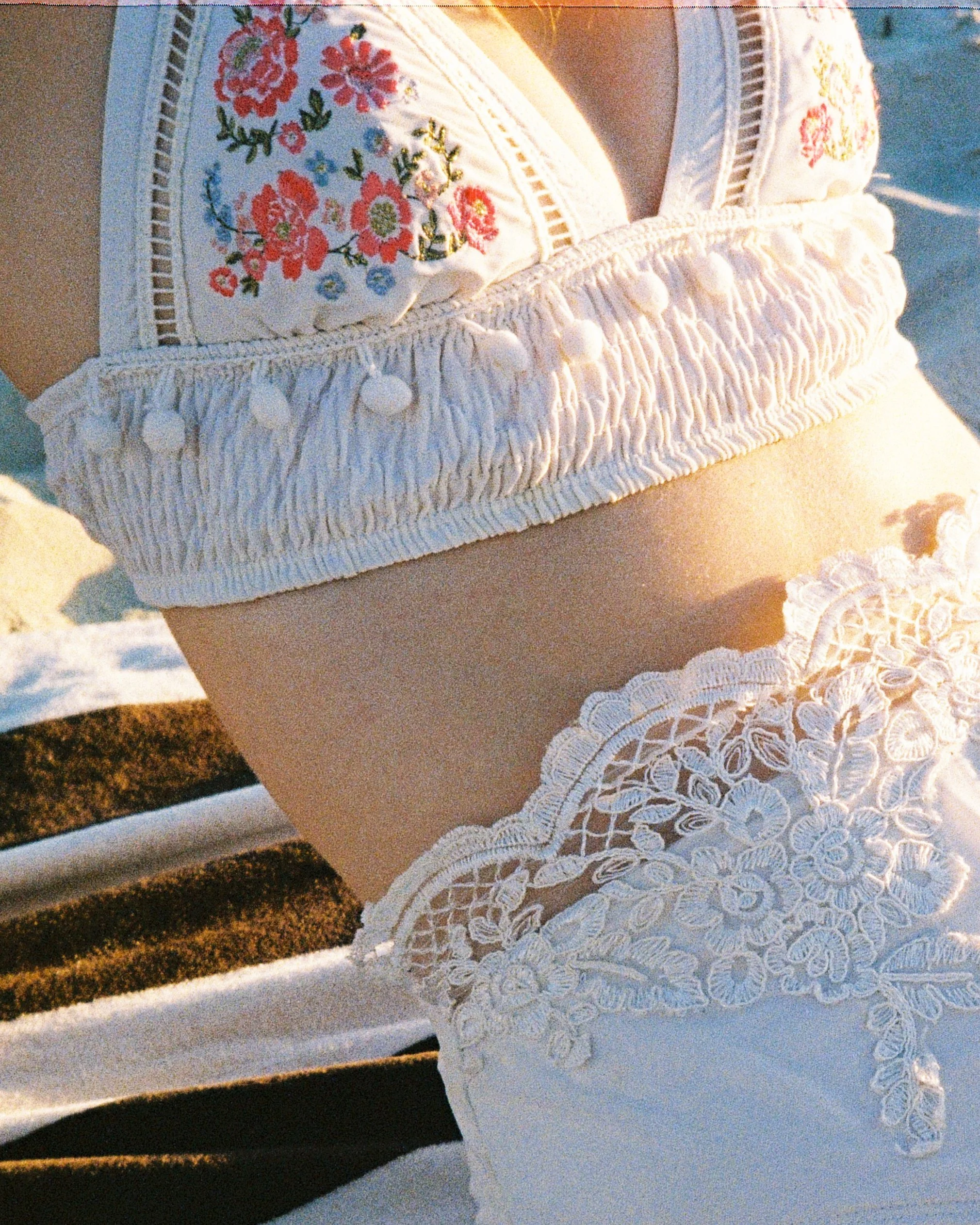 Close-up of a woman's waist and hips wearing white lace and embroidered swimsuit, sitting on a lounge chair by the beach during sunset.