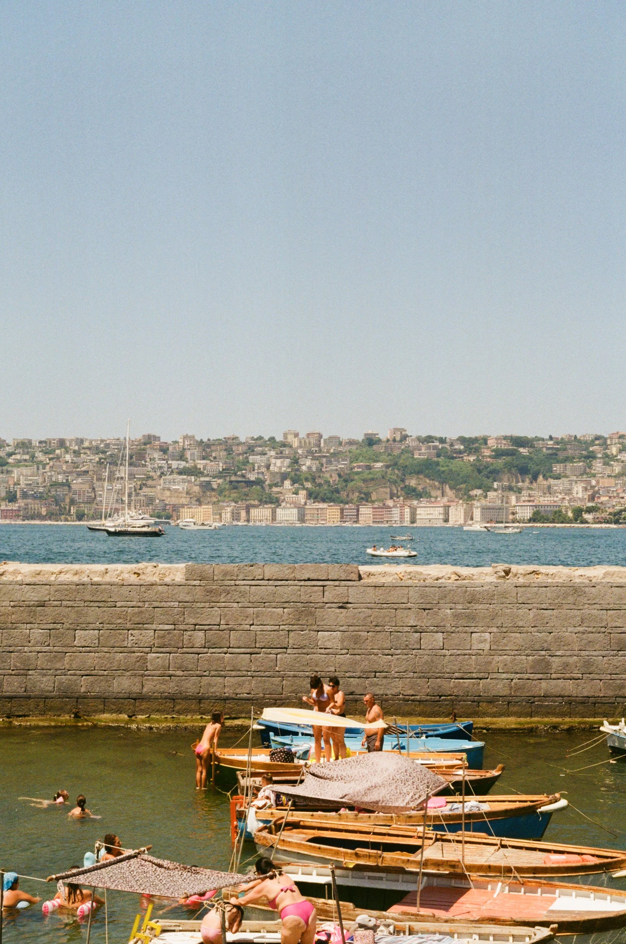 People swimming and relaxing near boats at a marina with a cityscape and hillside in the background.