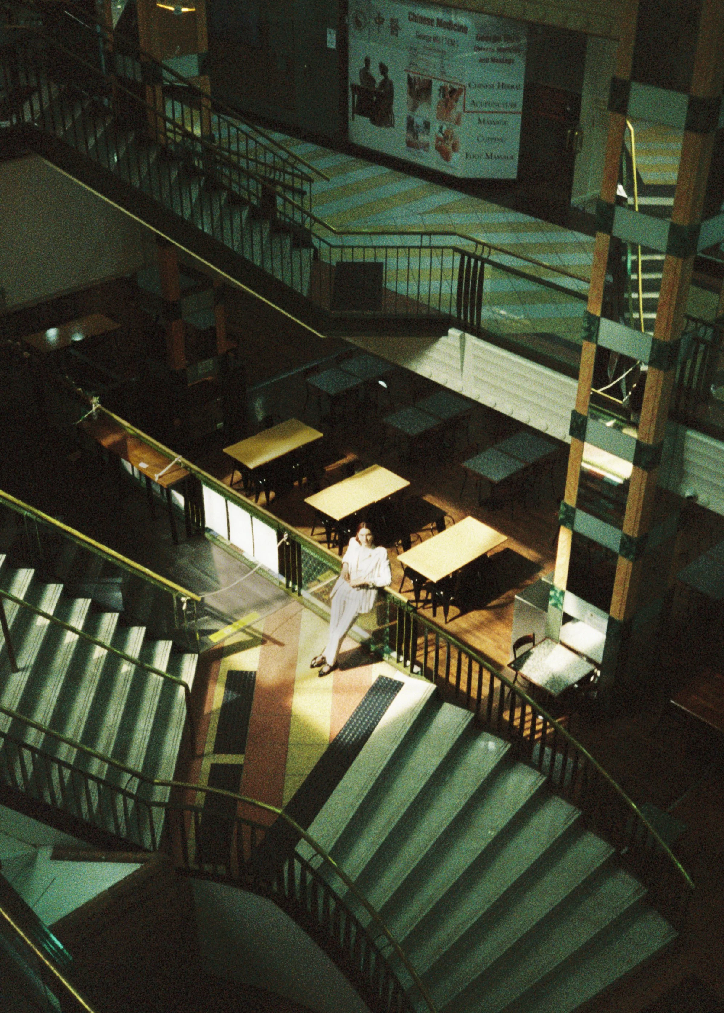 A woman standing on the stairs of a large, empty shopping mall, with a tiled floor, glass railings, and a staircase in the background. There is a sign for Chinese medicine on the wall.
