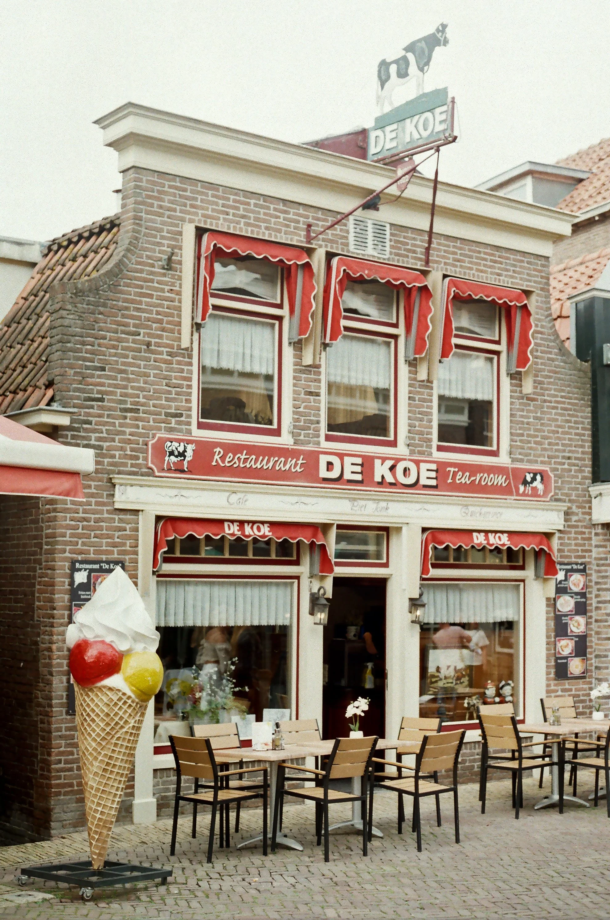 A brick restaurant with red awnings and signage reading De KOE, with outdoor seating and an ice cream cone statue outside.