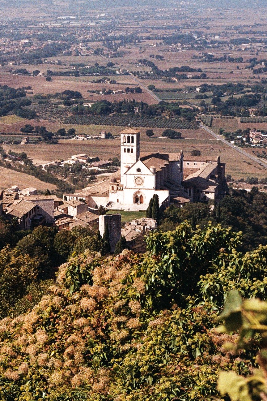 A historic church with a bell tower situated on a hillside, surrounded by trees and overlooking a landscape of rolling fields and farmland.