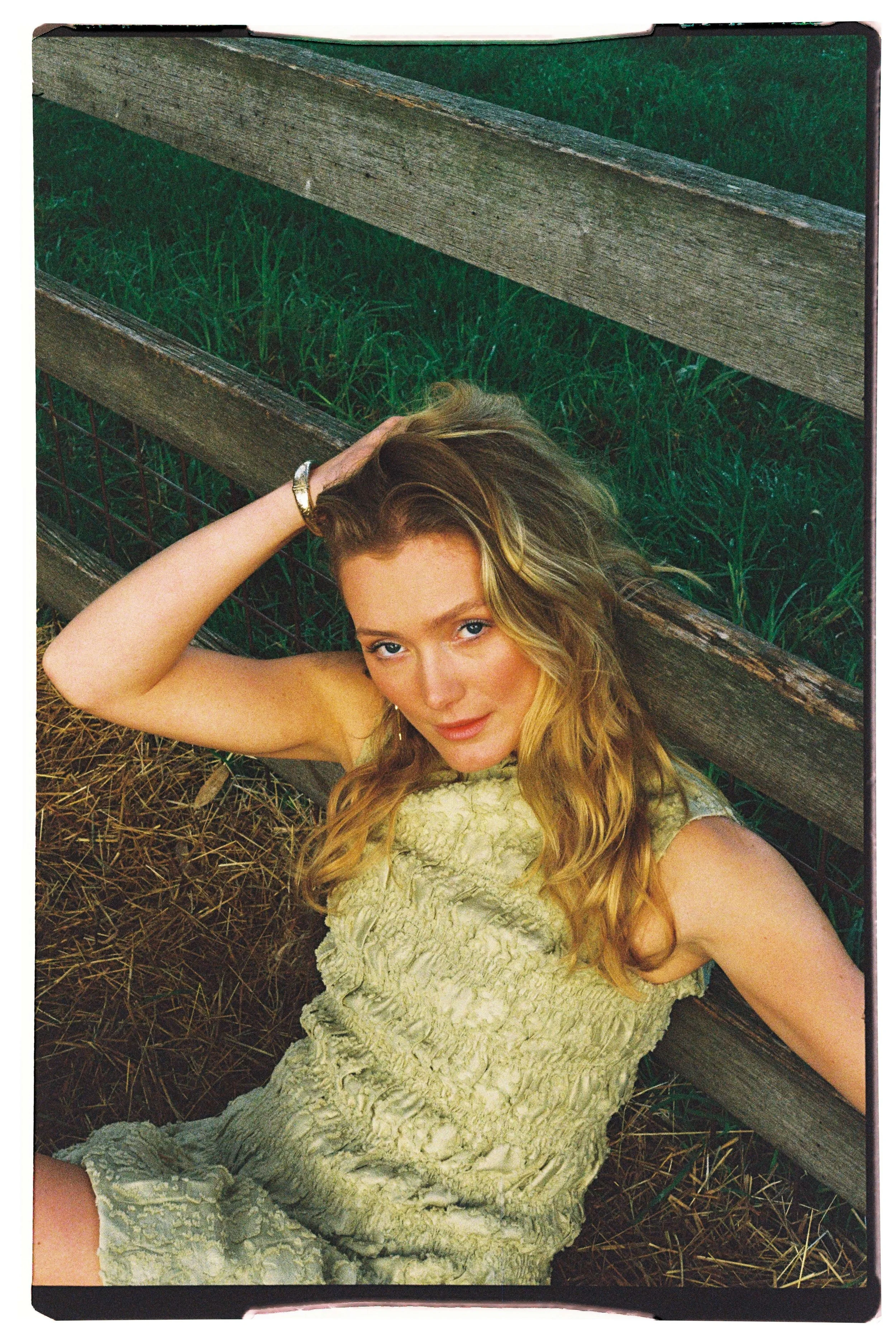 A woman with wavy blonde hair lying on the ground next to a wooden and wire fence, wearing a textured light-colored sleeveless dress, looking up at the camera with one hand on her head.