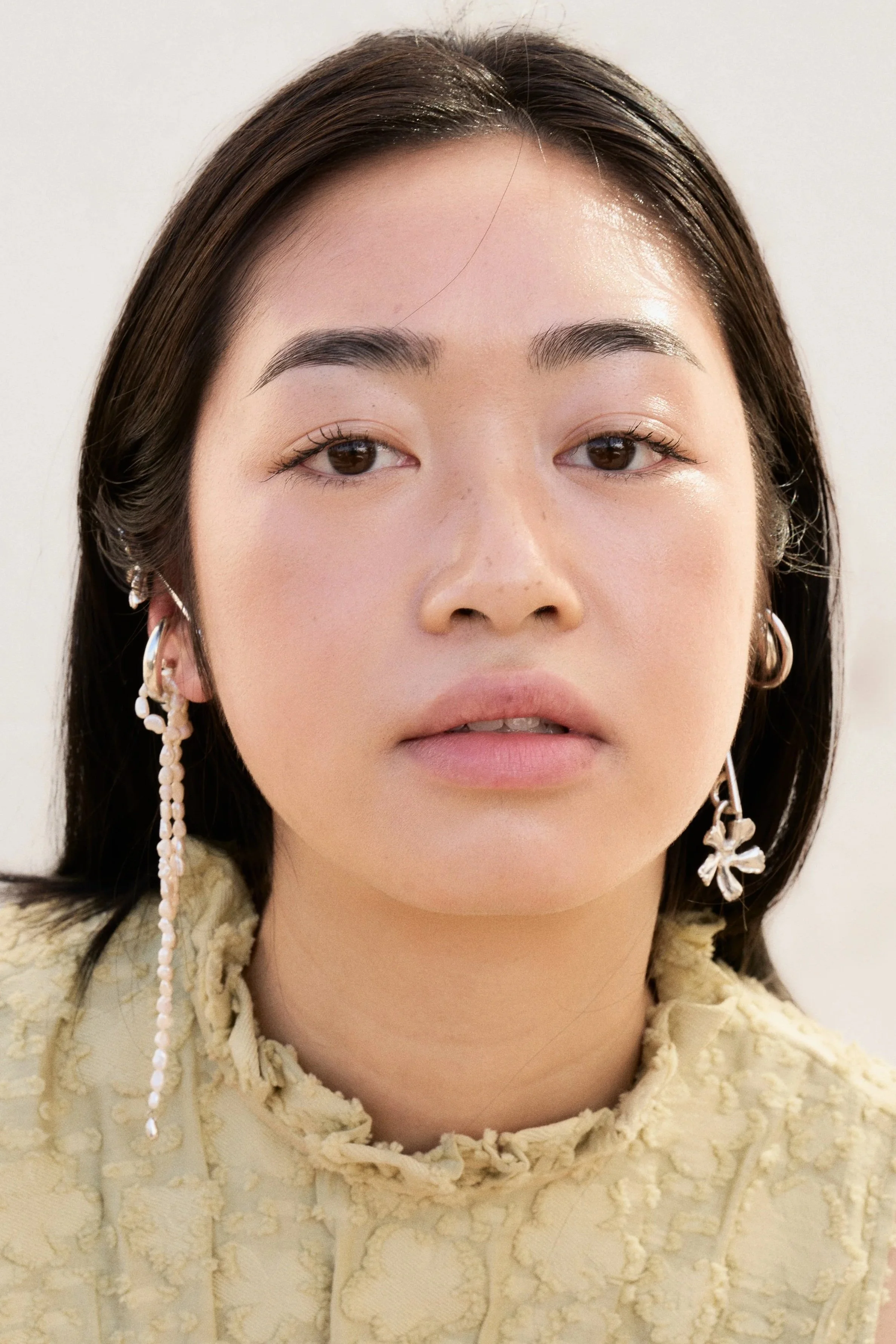 Close-up of a young woman with dark hair and makeup, wearing a cream-colored textured top and jewelry including earrings with pearl details and a flower design.