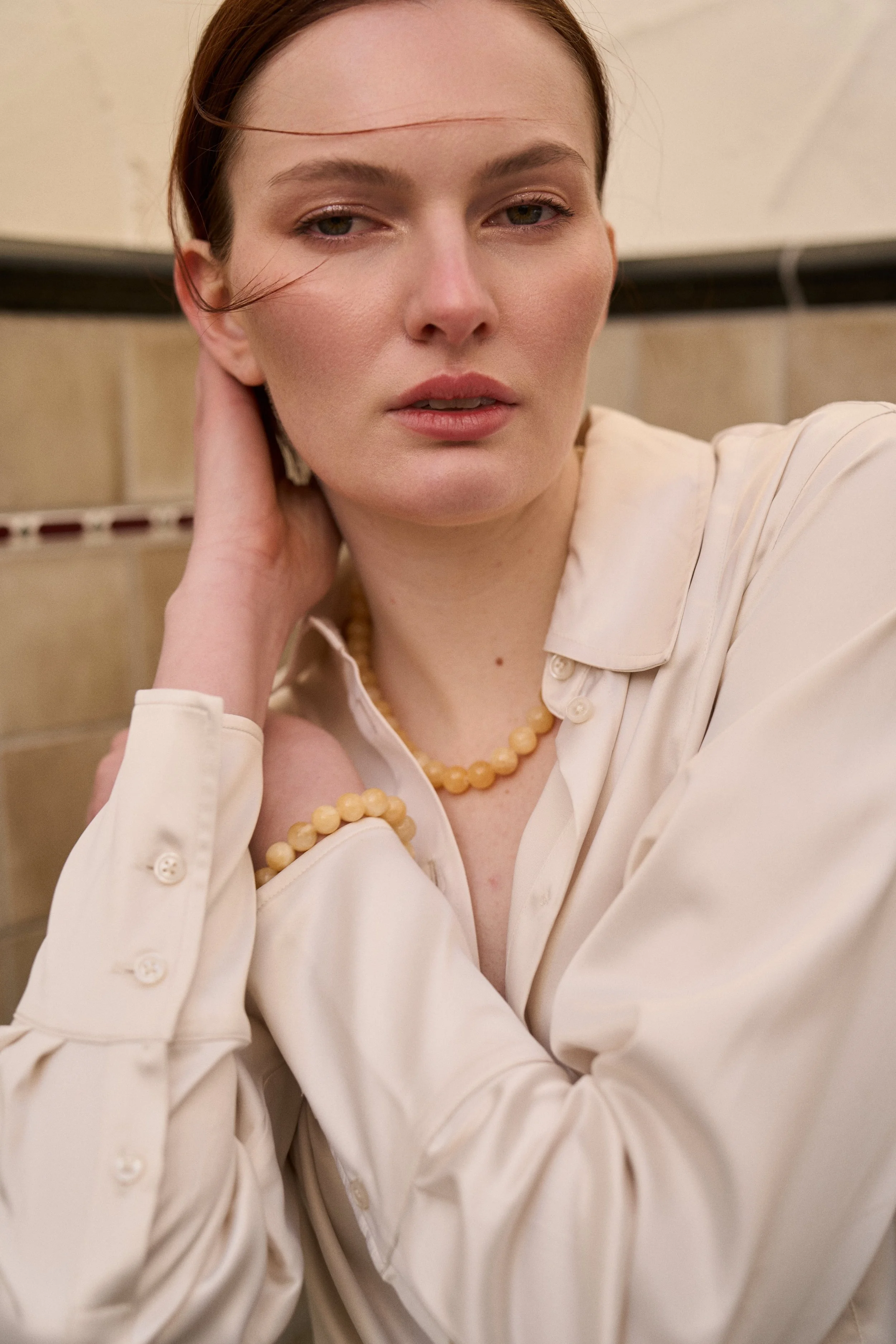 A woman with red hair and fair skin wearing a cream-colored blouse and a pearl necklace, posing indoors against a tiled wall.