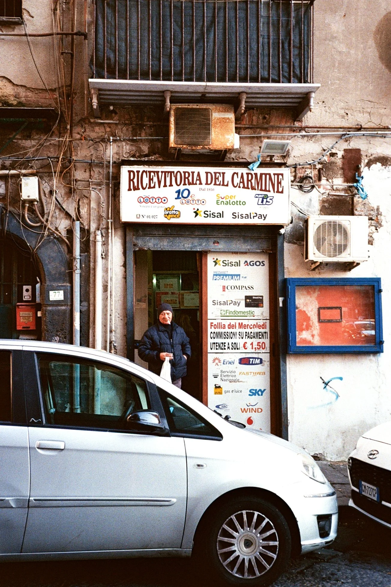 A small store called 'Ricevitoria del Carmine' on a narrow street, with a man standing at the entrance, surrounded by parked cars and various electrical wires on the wall.