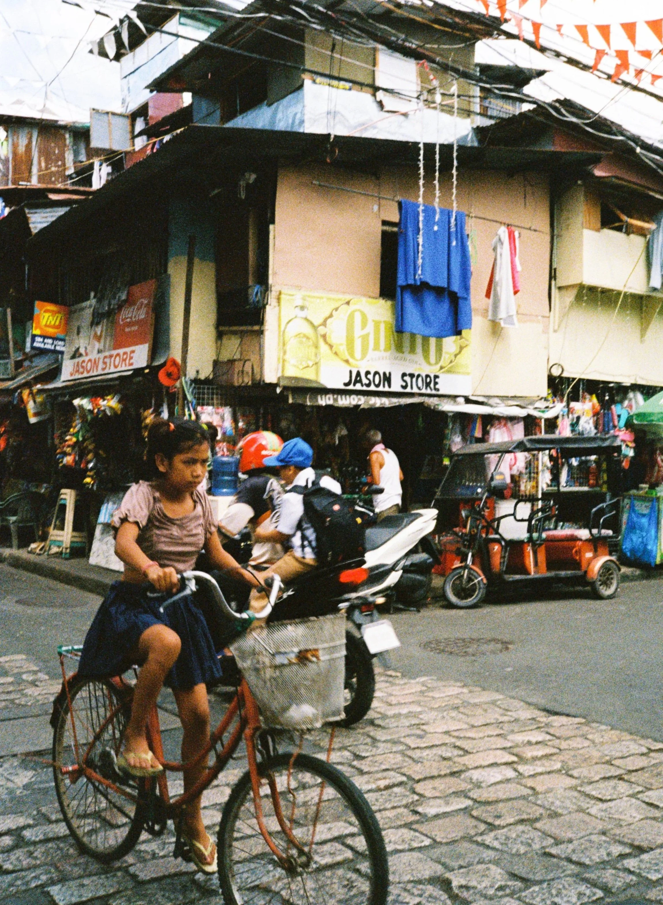A young girl riding a bicycle on a cobblestone street in a busy market area. There are shops with signs, motorcycles, and tricycles parked along the street. Several people are walking or on motorbikes, and laundry hangs above the shops.