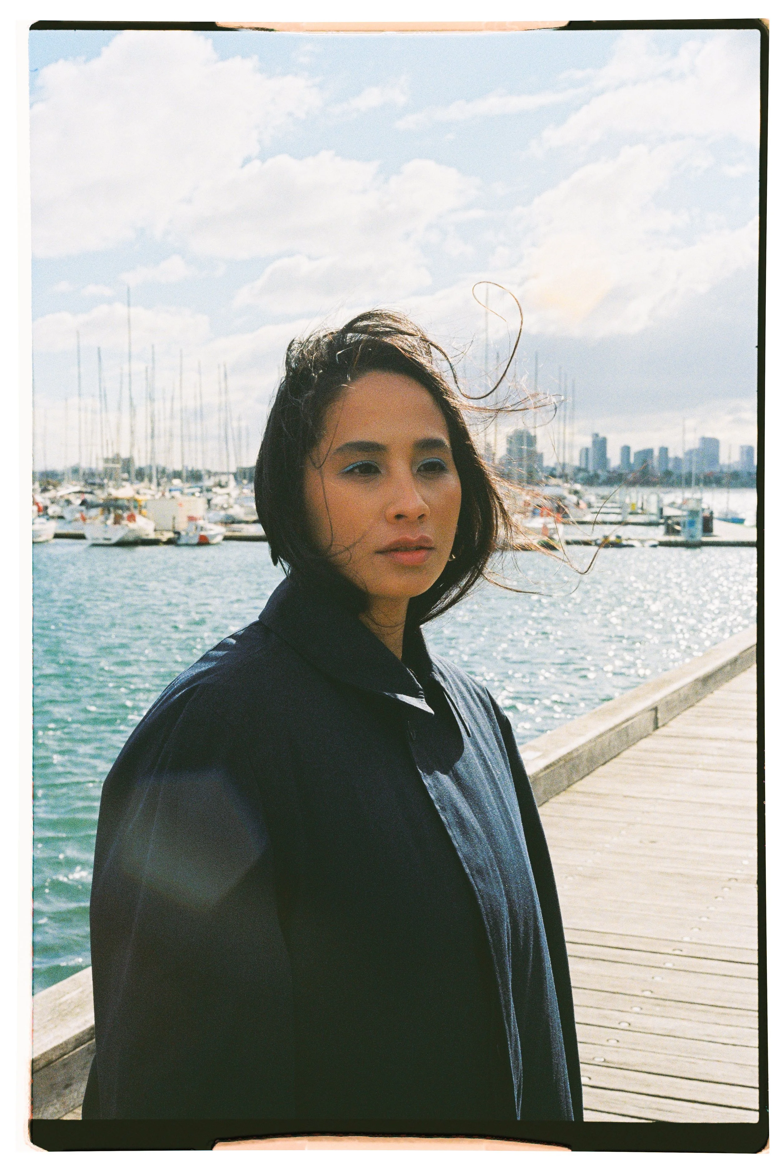 A woman with shoulder-length dark hair and blue eyeshadow standing on a wooden pier by a marina with boats and a city skyline in the background, with the wind blowing her hair.