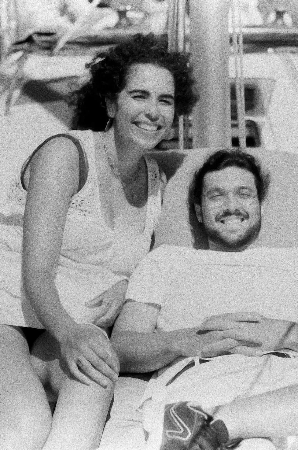 A woman with curly hair smiling poses with a man lying in a hospital bed, also smiling. The scene appears to be outdoors near a pool or beach area.