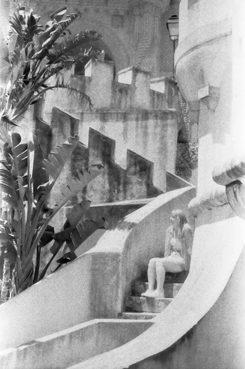 A woman sitting on a curved staircase, surrounded by large potted plants and architectural features such as walls, a lamp, and a balcony.