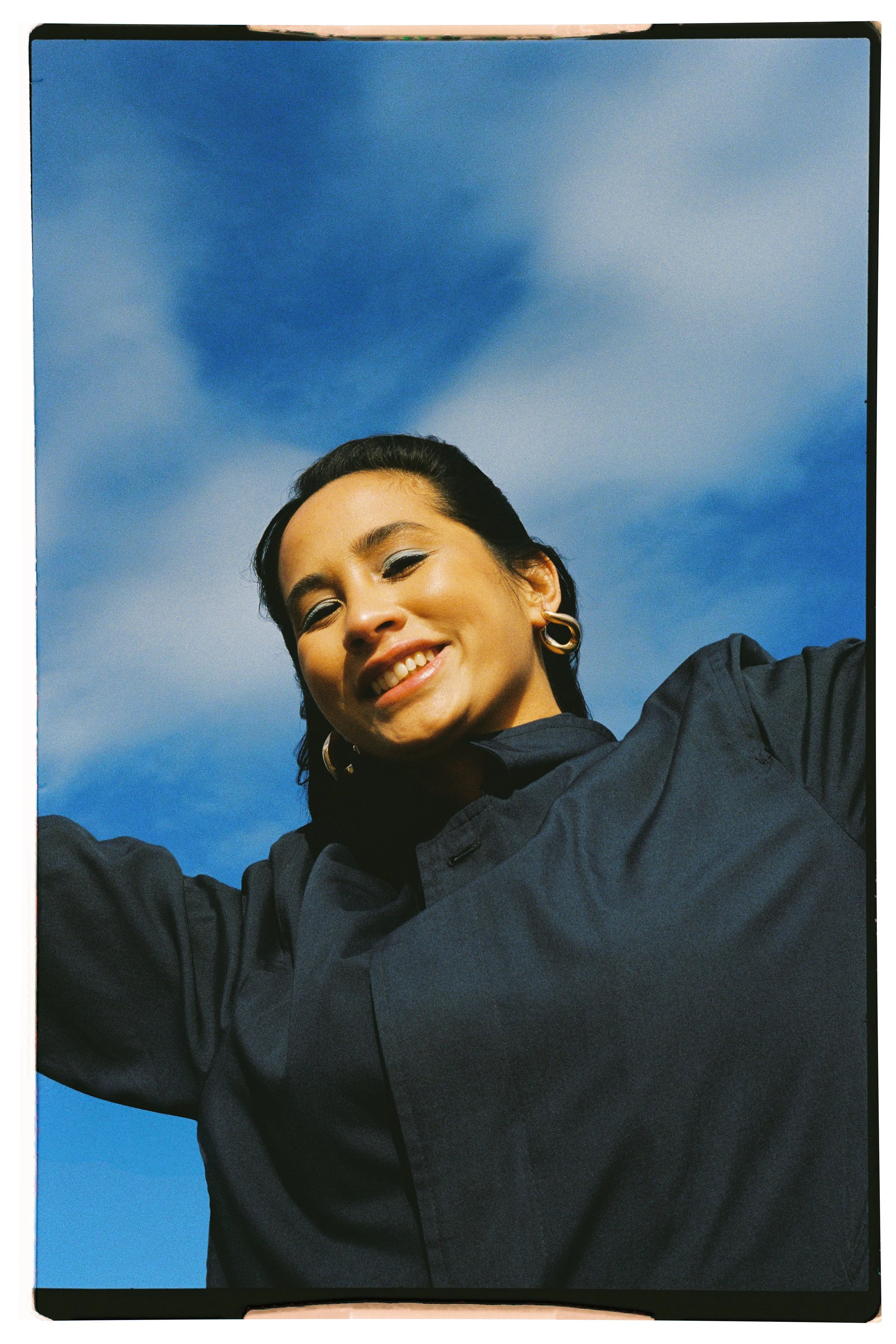 A woman smiling outdoors against a blue sky with clouds, wearing a dark shirt and hoop earrings.