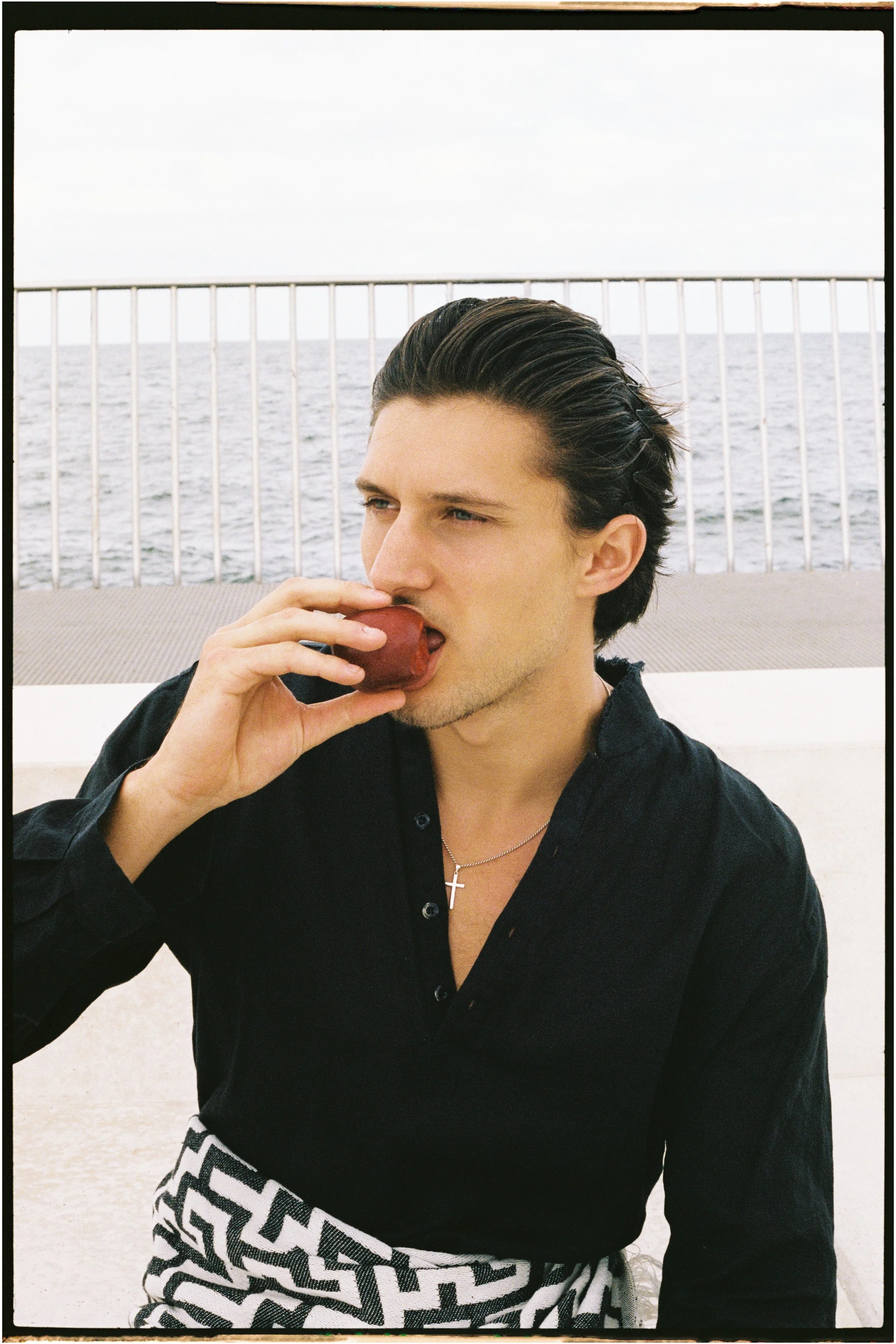 Man with dark hair, wearing black shirt and a silver cross necklace, eating an apple near the ocean on a deck with a metal railing.