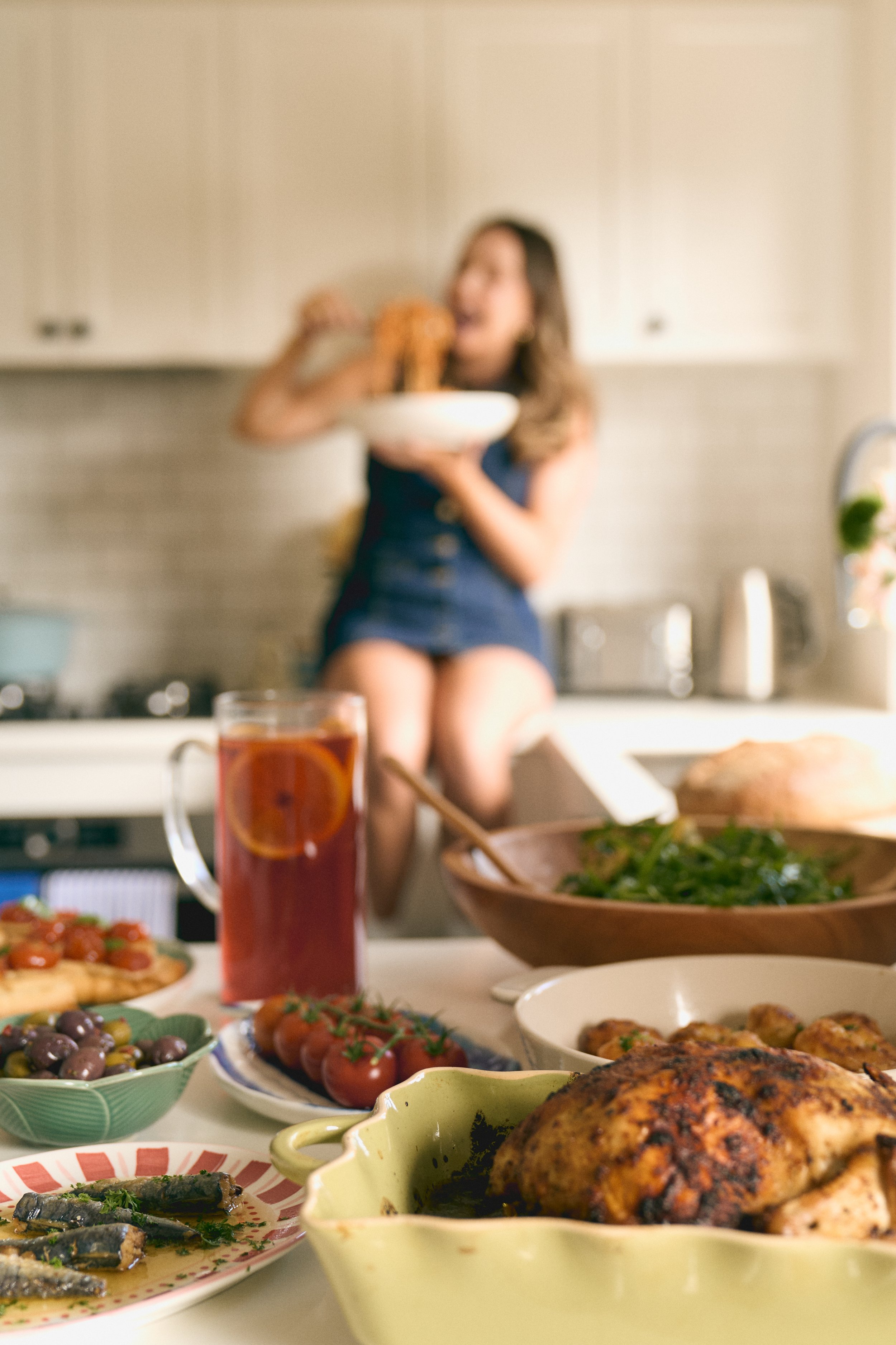 A woman sitting on a kitchen counter in the background, holding a bowl and eating food. The foreground shows a table filled with various dishes, including a roasted chicken, salad, tomatoes, olives, a drink with slices of orange, and other plated foods.