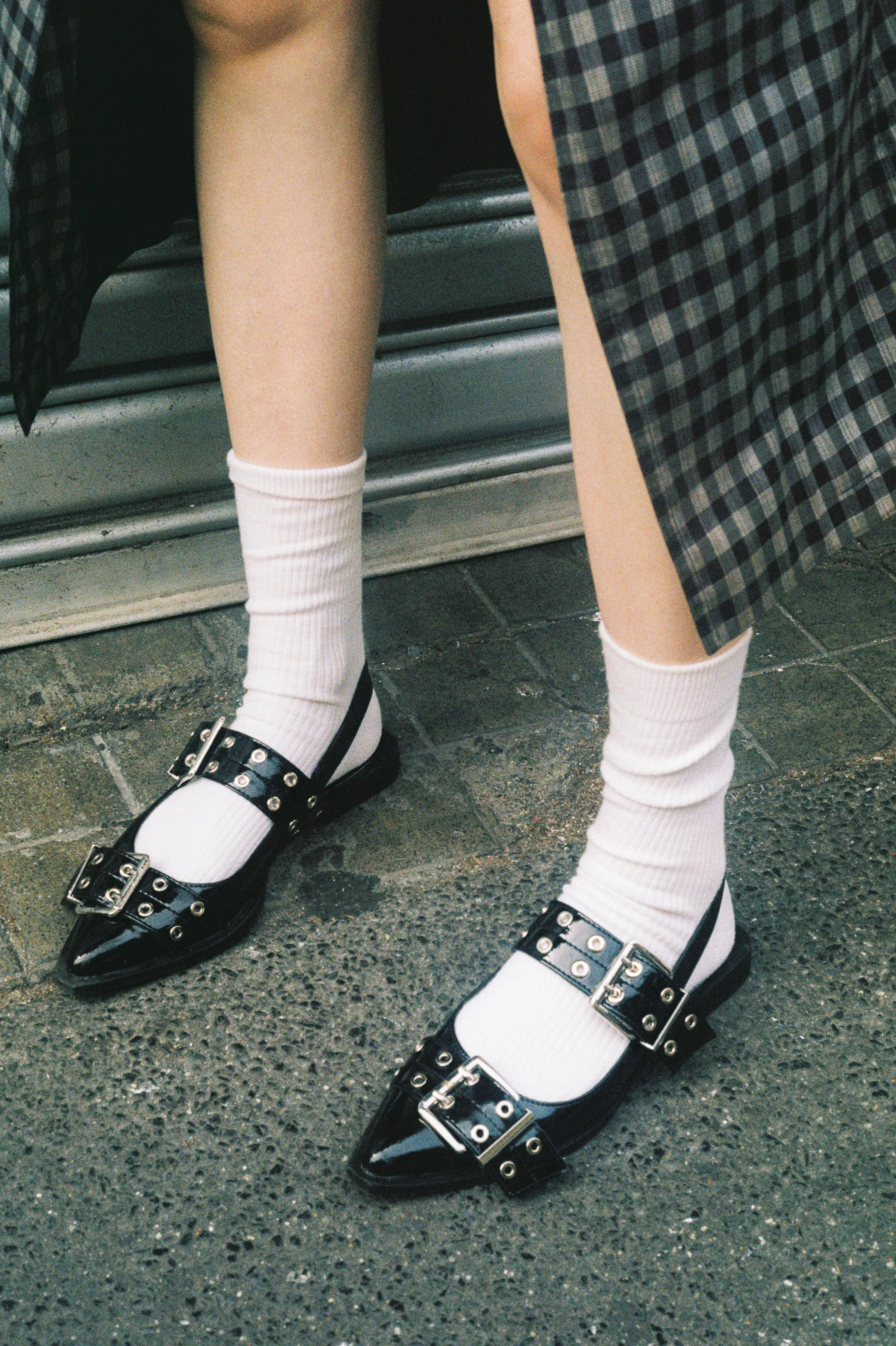 Close-up of a person's legs wearing white socks, black pointed flats with buckles and studs, a checkered skirt, and standing on a textured asphalt pavement near a metallic surface.
