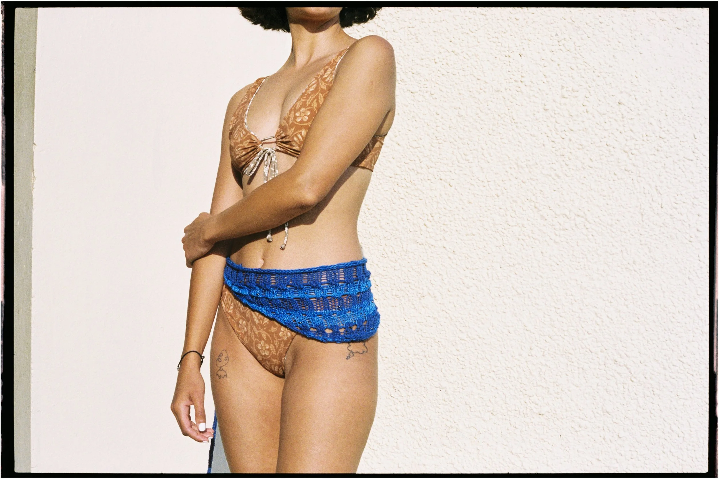 A woman with curly dark hair wearing a semi-transparent floral bikini top, lace bikini bottom, and a blue crocheted wrap around her waist, standing against a plain light-colored textured wall.