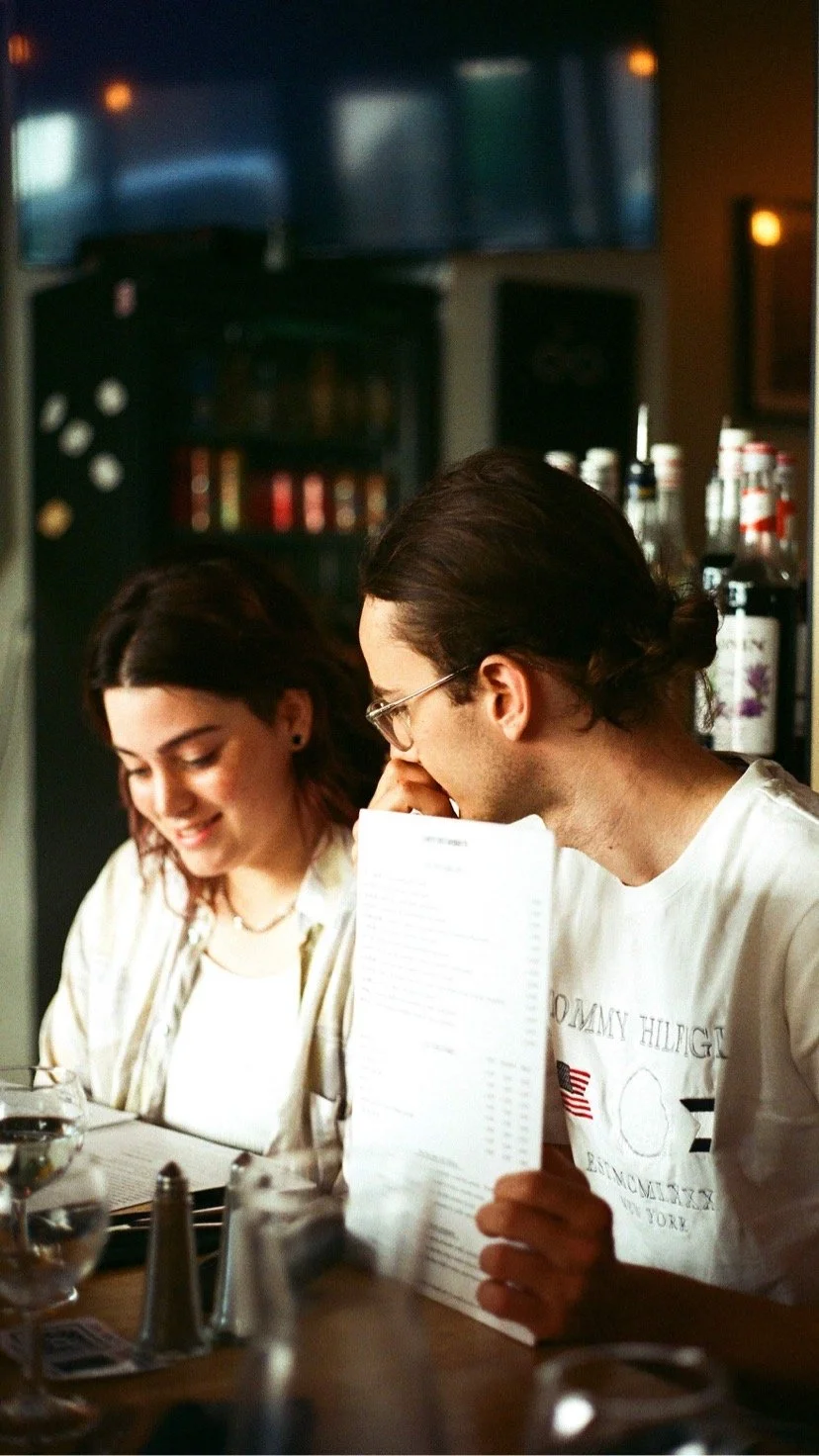 A man and woman sitting at a table in a restaurant, looking at a menus, with bottles of condiments visible in the background.